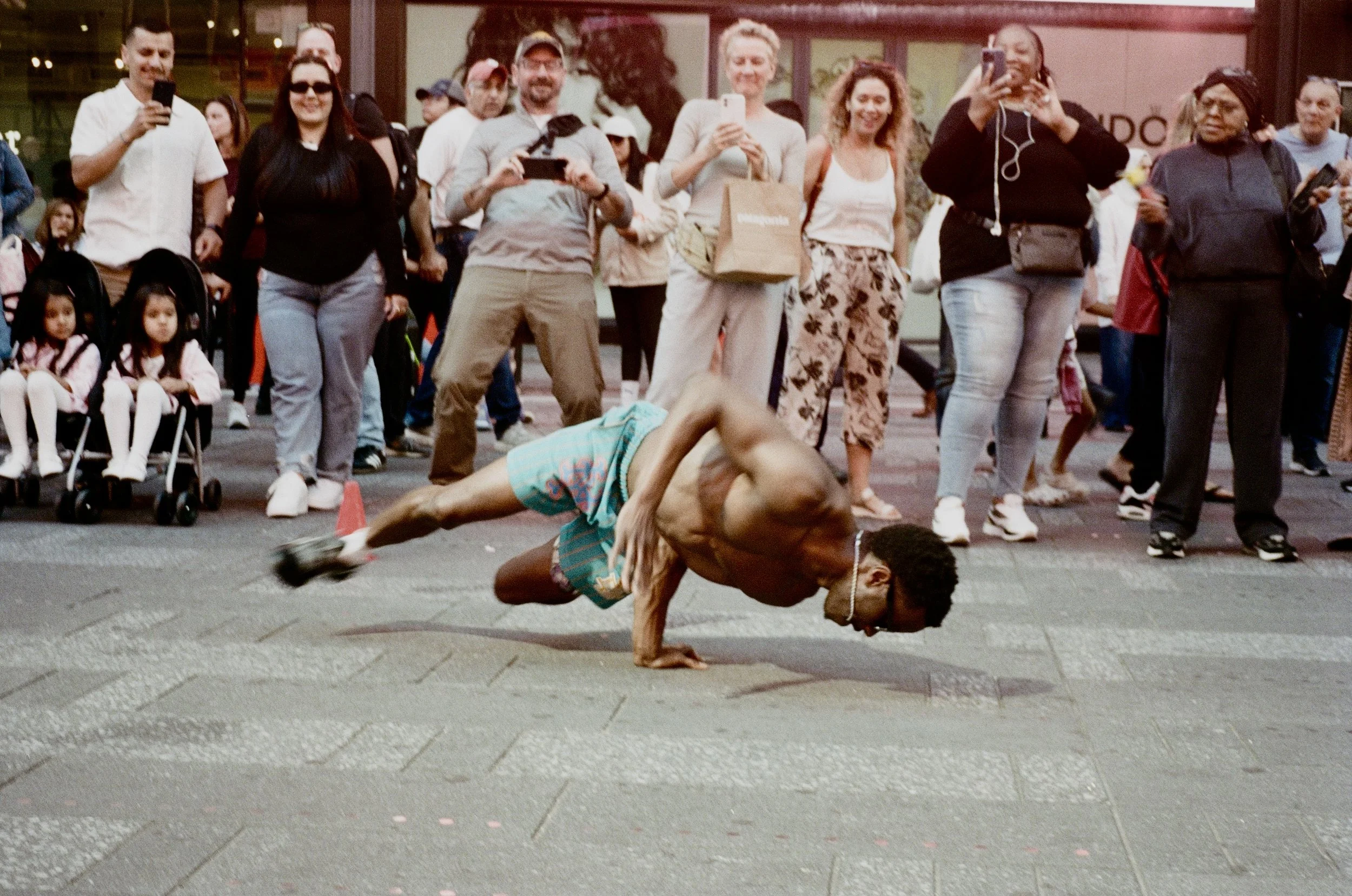 A shirtless man with glasses performing a breakdance move on the sidewalk, surrounded by a crowd of onlookers taking photos and watching him.