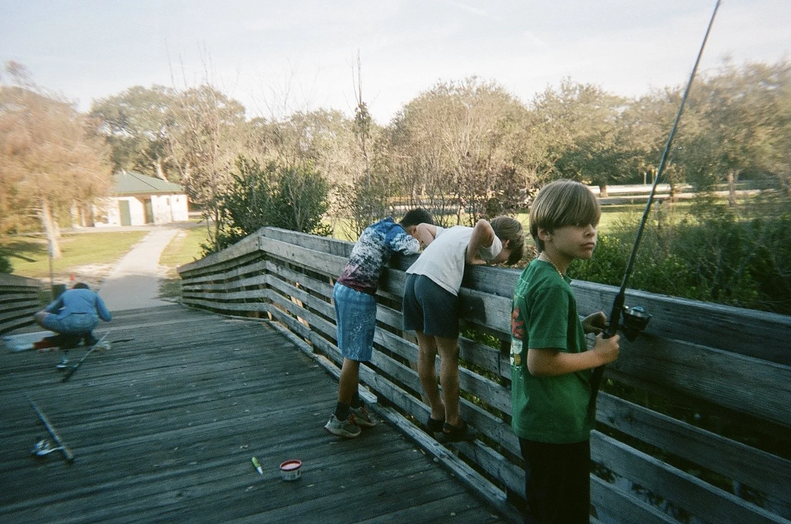 Group of children on a wooden bridge, one boy in a green shirt holding a fishing rod, other children looking over the railing, in a park with trees and a trail in the background.