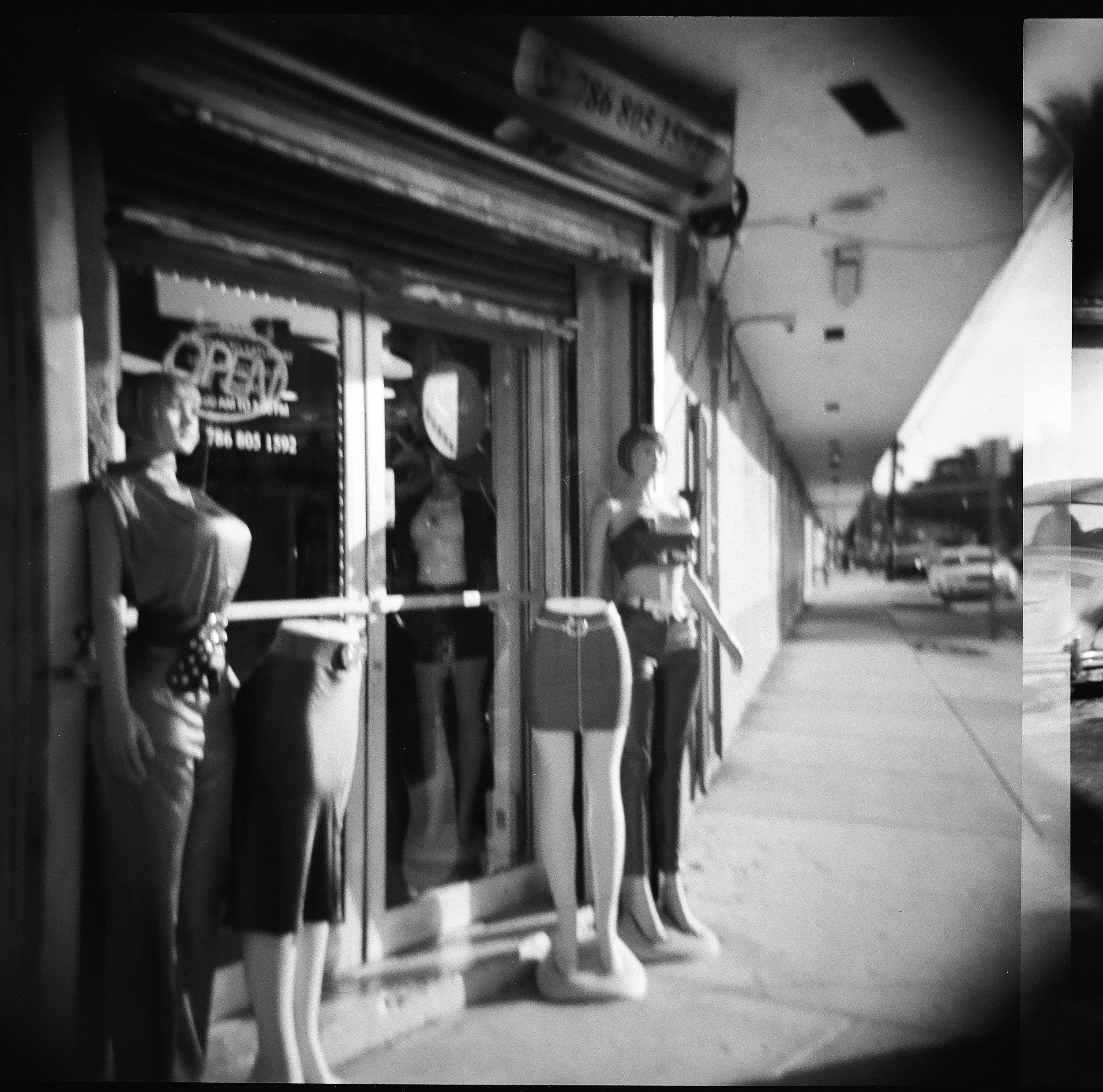 Black and white photo of store mannequins outside a clothing store on a sidewalk, with cars parked on the street in the background.