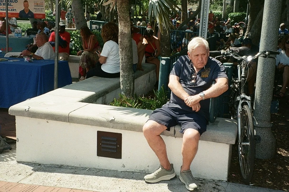 An elderly man with white hair sitting on a concrete bench in a park, leaning against a tree with his arms crossed. He is wearing a dark short-sleeved shirt, shorts, and sneakers, and has sunglasses hanging from his shirt collar. A bicycle is parked 