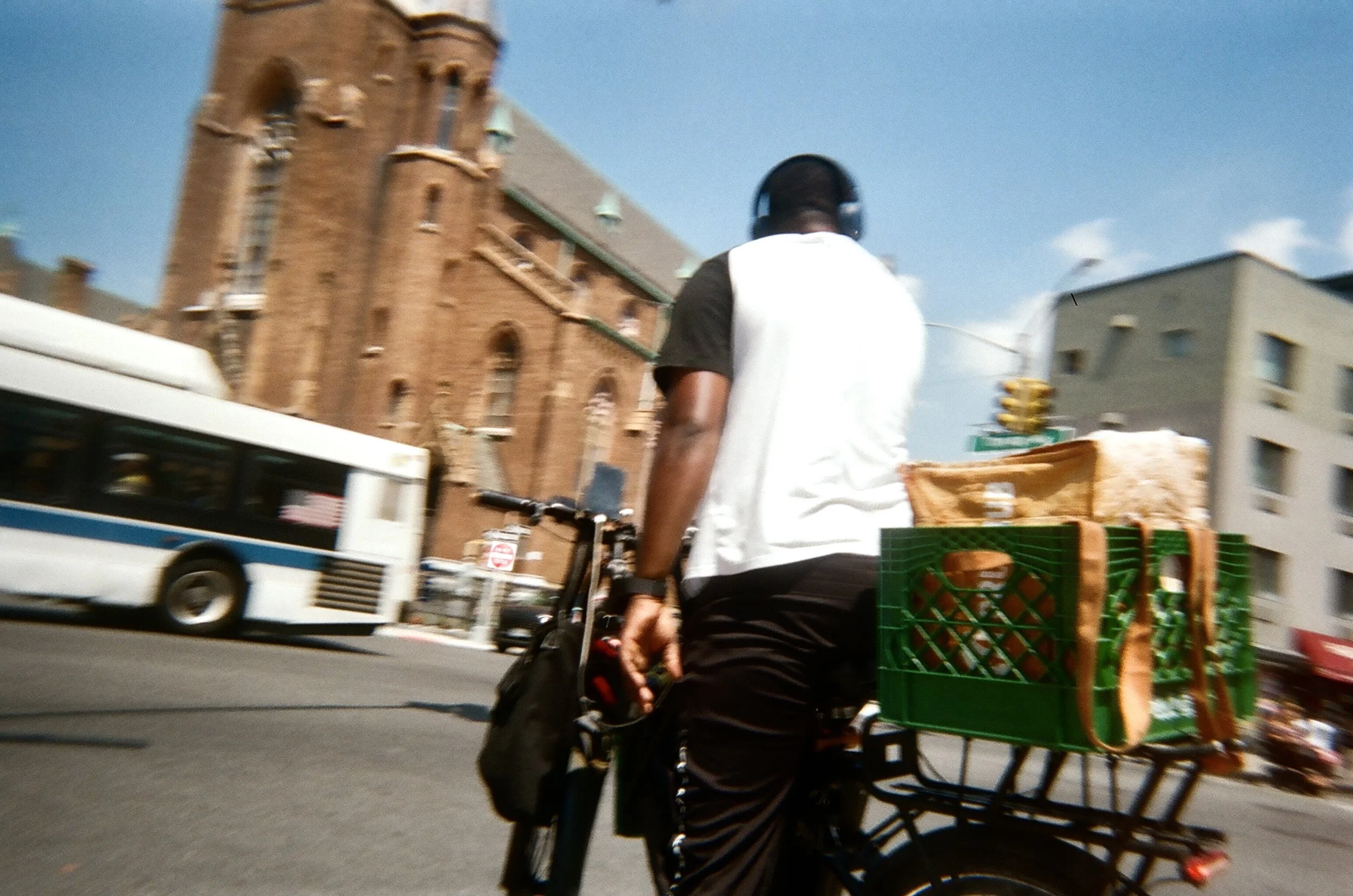 A man riding a bicycle on an urban street with a large green crate containing bread. A white bus and multi-story buildings are visible in the background.