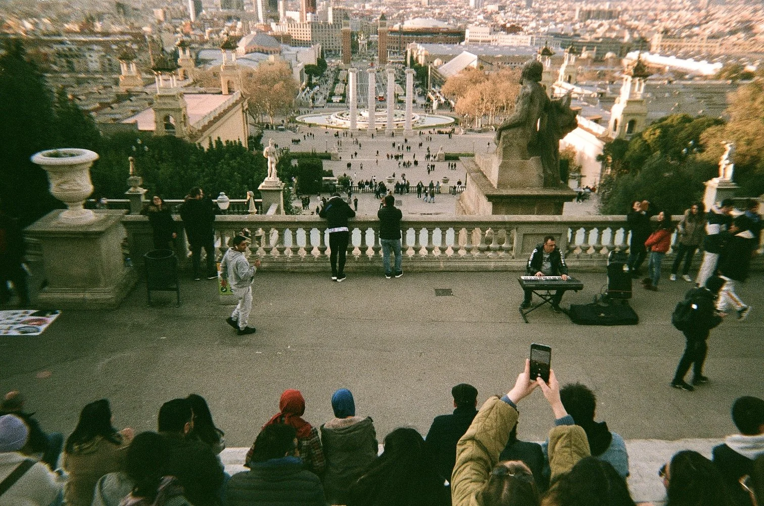 People sitting on steps overlooking a large square with statues and trees, and a cityscape in the background.