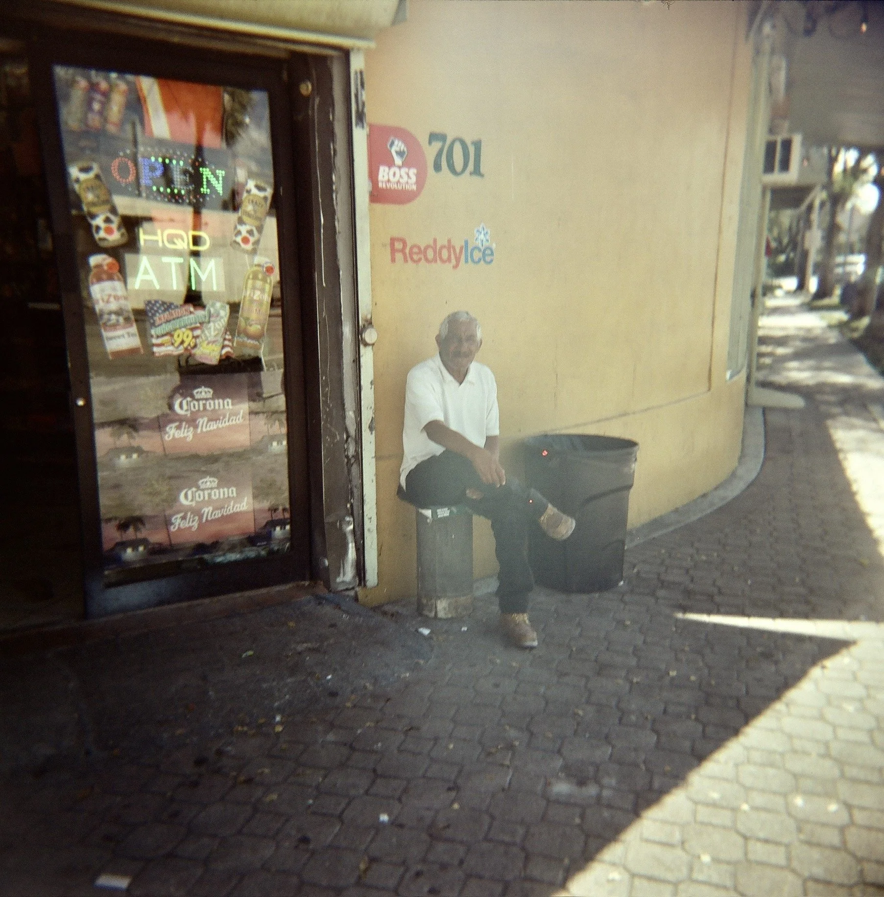 Older man sitting on a bench outside a store, wearing a white shirt and dark pants, with a trash can nearby.