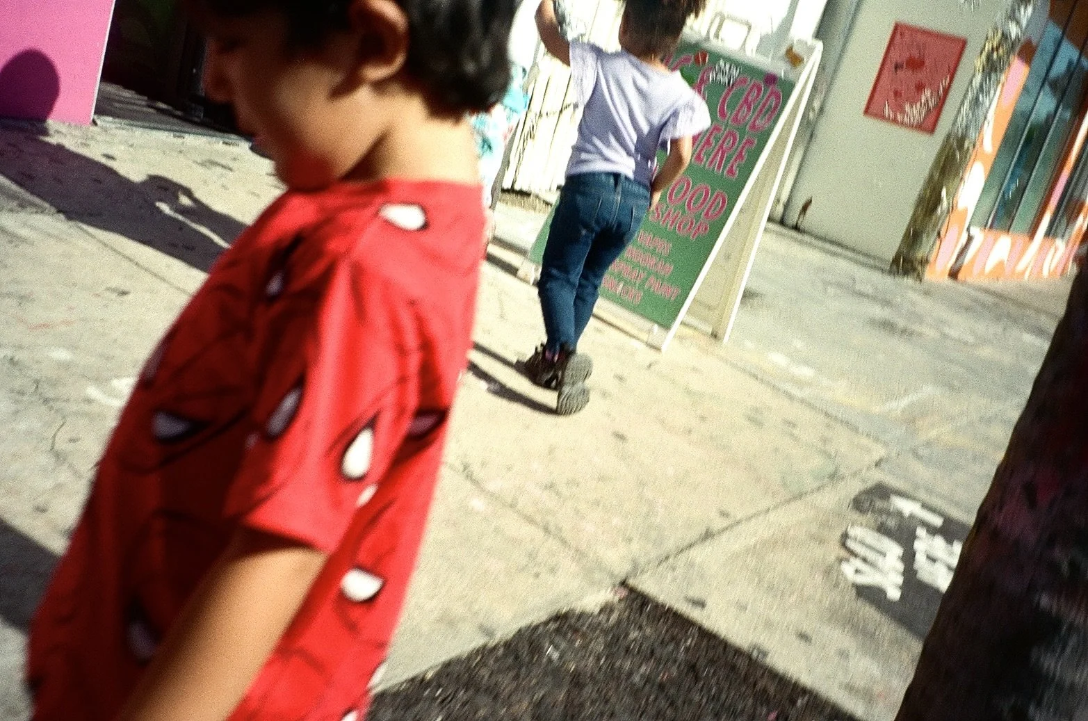A young child in a red shirt with Spider-Man mask print walking on a sidewalk, another child in a purple top and jeans standing near a green sidewalk sign for a food shop, and a storefront with colorful signage and a window behind.