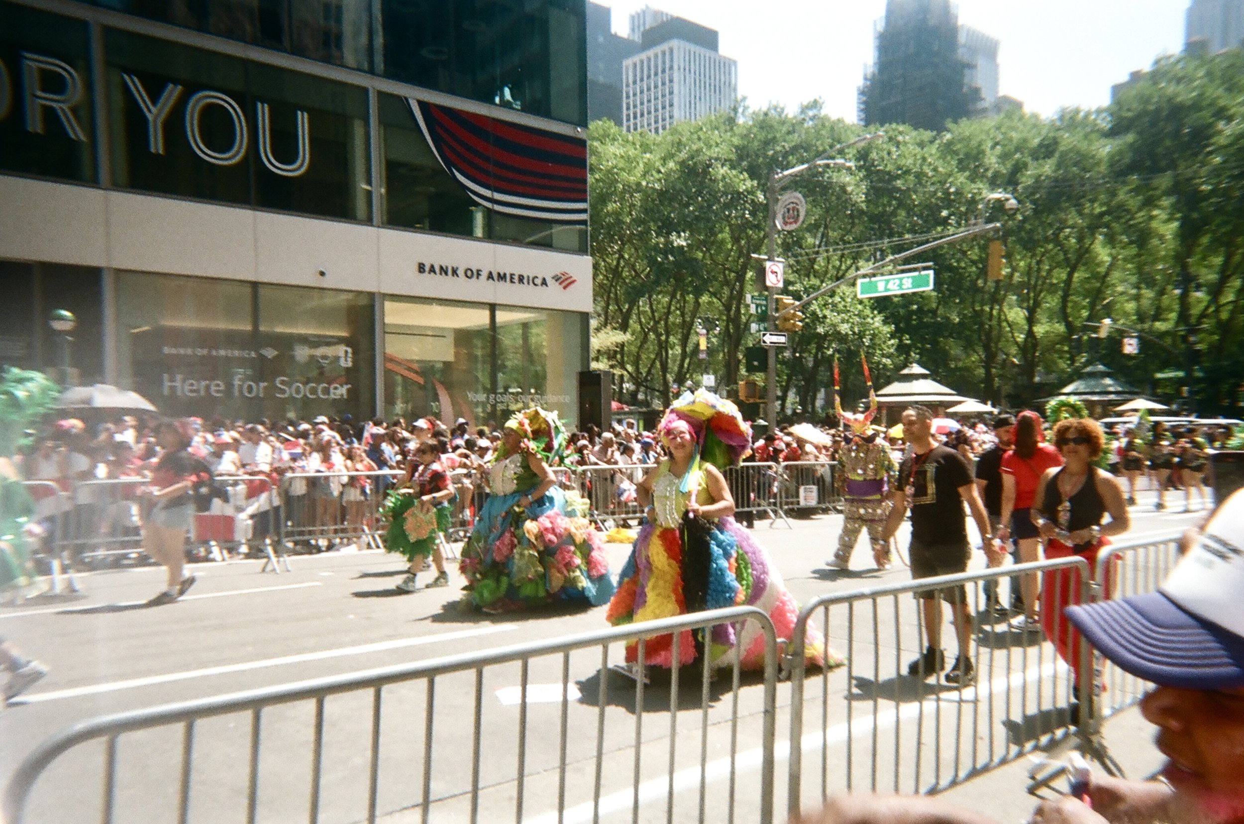 Colorful parade with people in vibrant costumes, some with large floral headdresses, walking along a city street lined with metal barricades, with a crowd of spectators behind the barricades, and tall buildings and trees in the background.