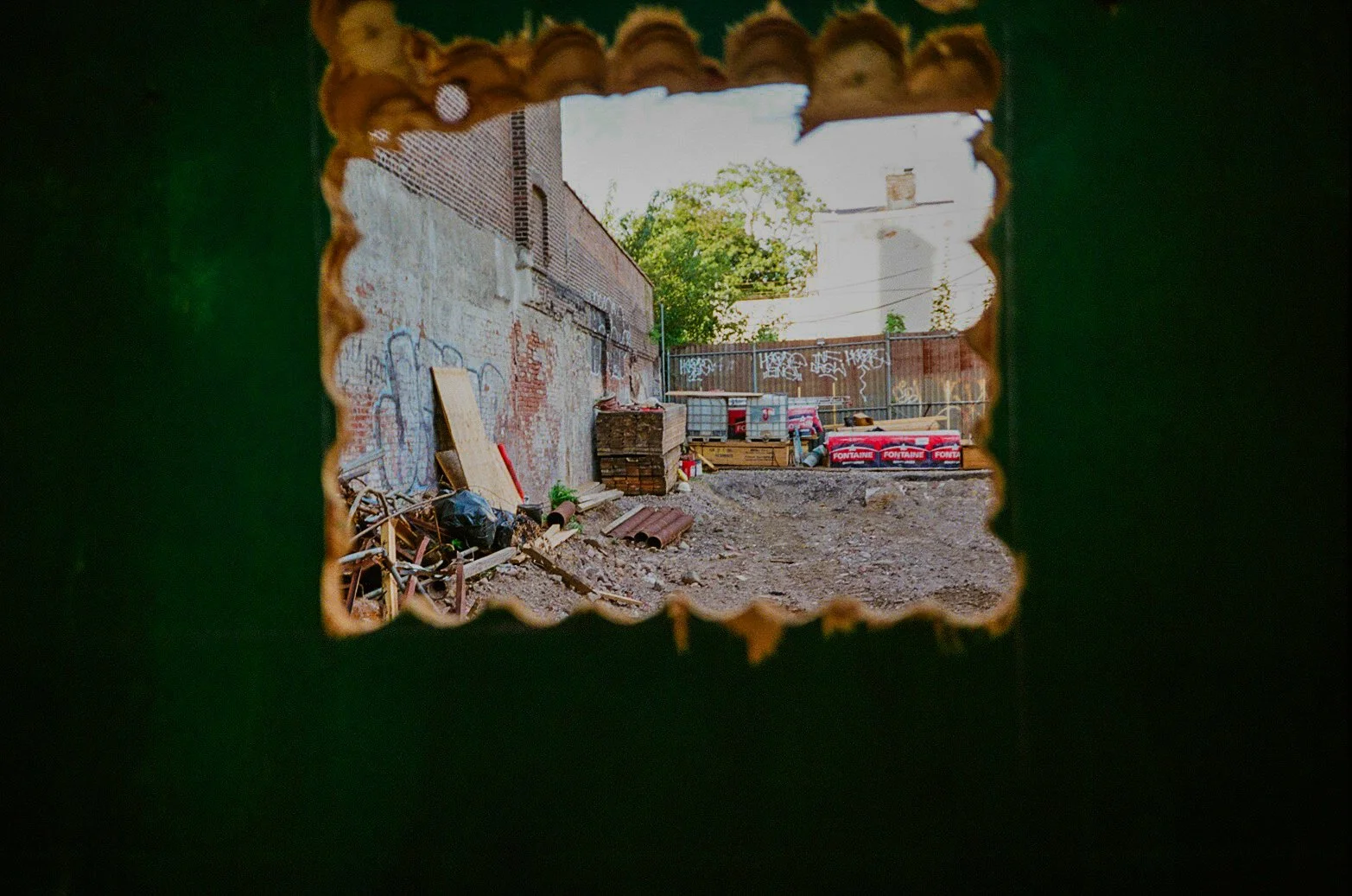 View of a messy construction or renovation site through a jagged hole in a green wall, showing debris, bricks, and construction materials against a brick wall with graffiti.