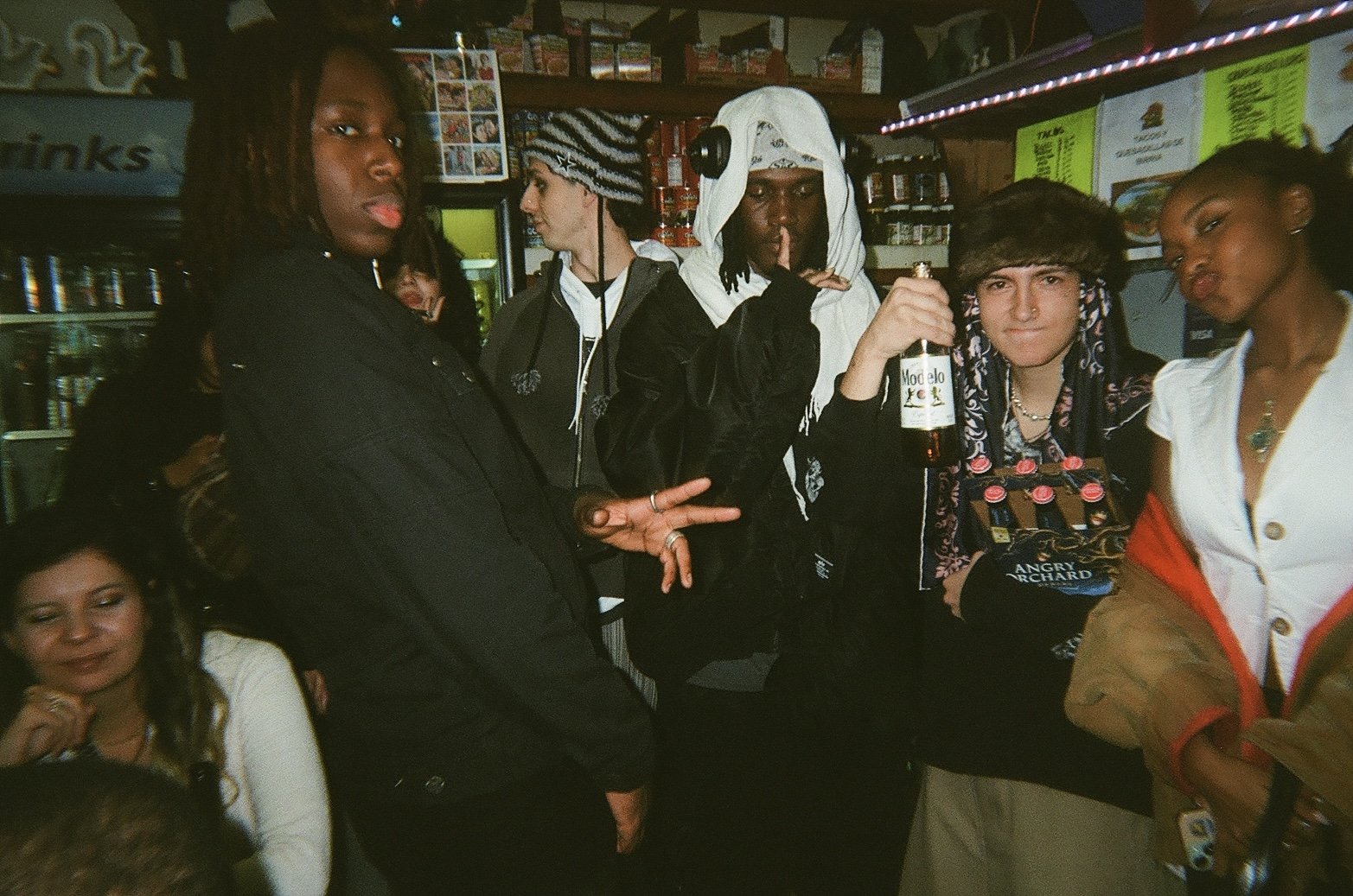 Group of young people socializing in a bar or club, some holding drinks, wearing casual and trendy clothing, with shelves of bottles in the background.