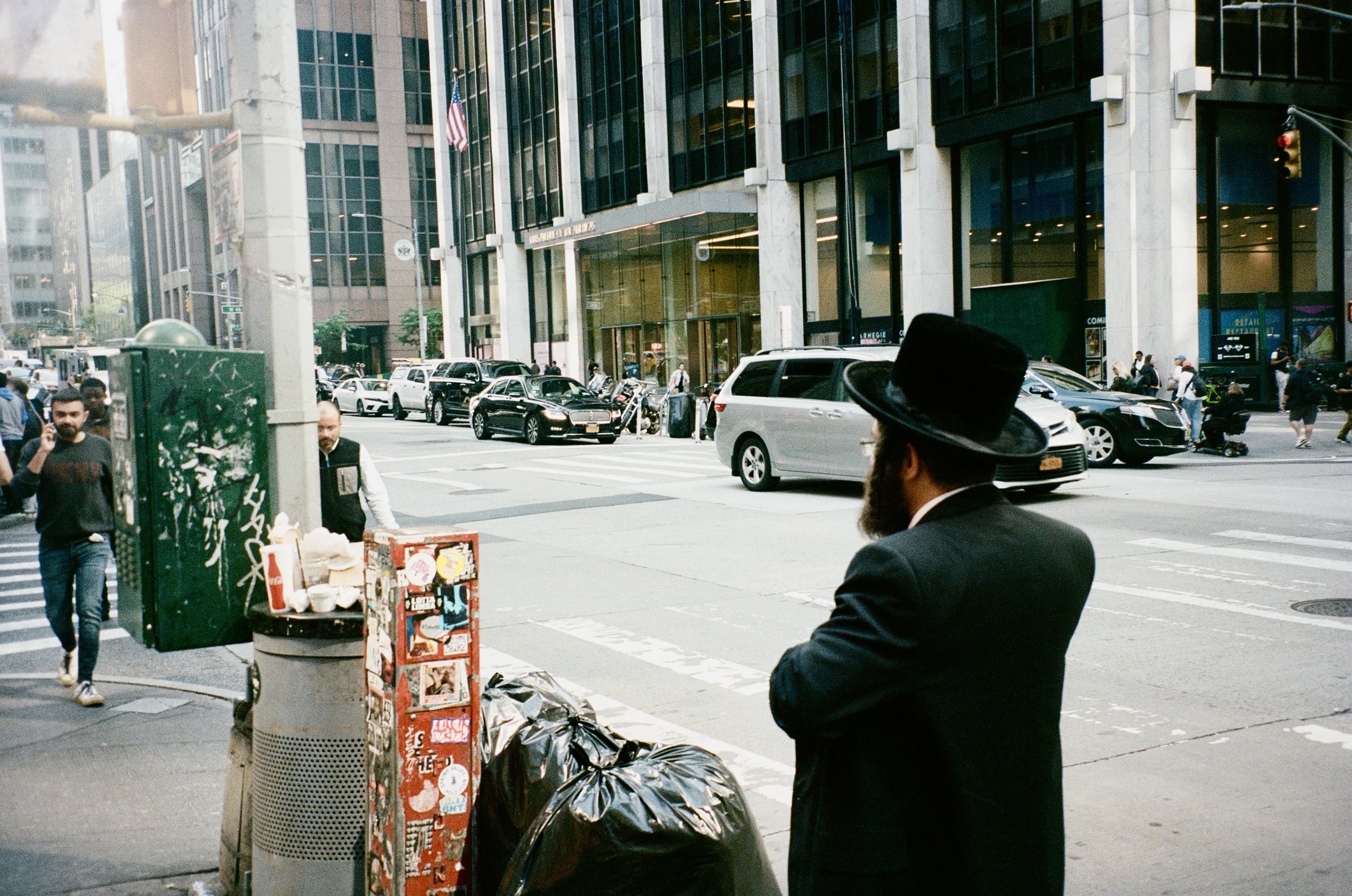 A man in a black suit and hat stands on a city sidewalk looking toward the street. There are cars, pedestrians, and tall office buildings in the background.
