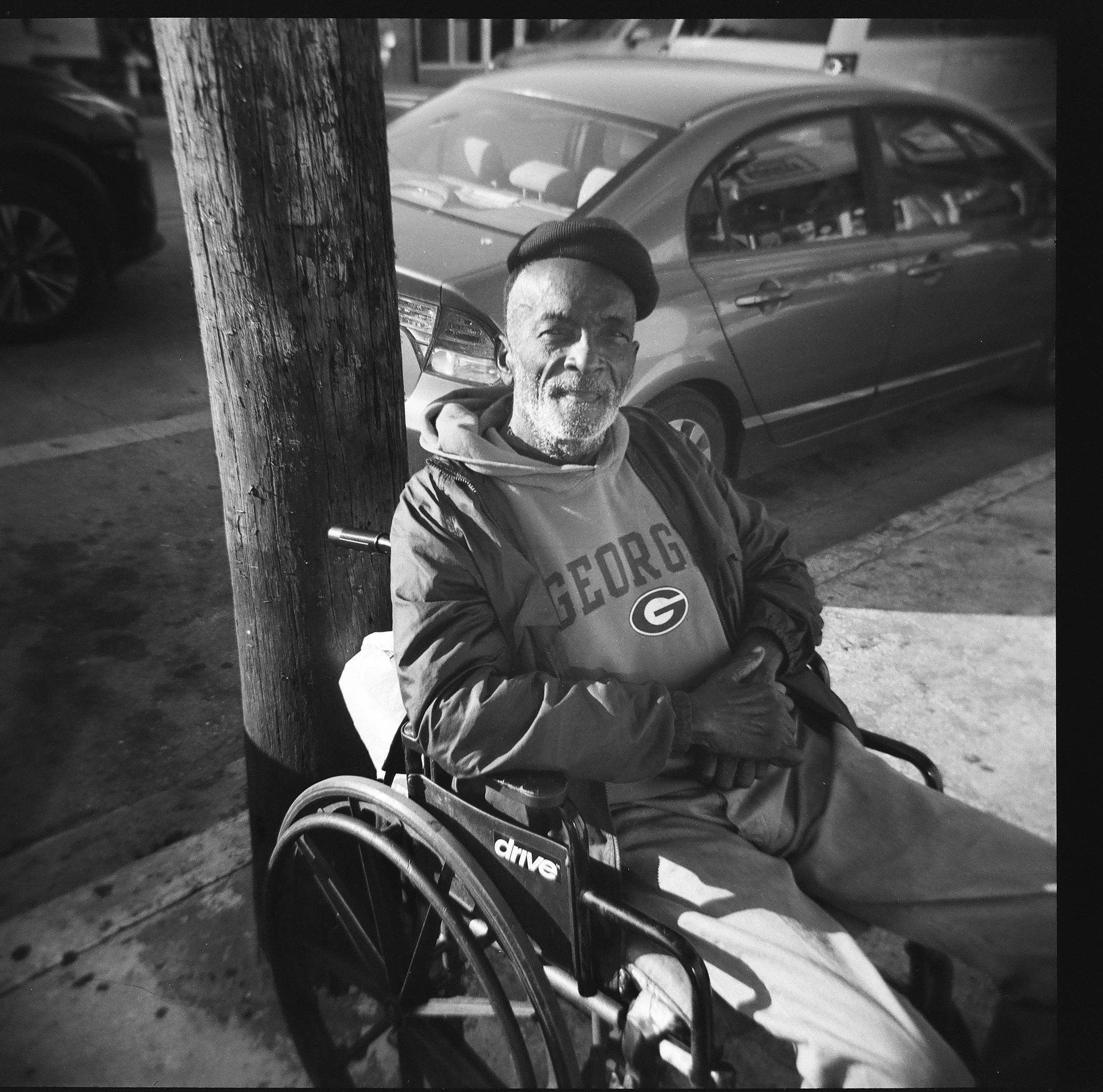 An elderly man with a beard and a hat, sitting in a wheelchair beside a wooden utility pole on a city street, wearing a Georgia Bulldogs sweatshirt and a jacket, smiling at the camera.