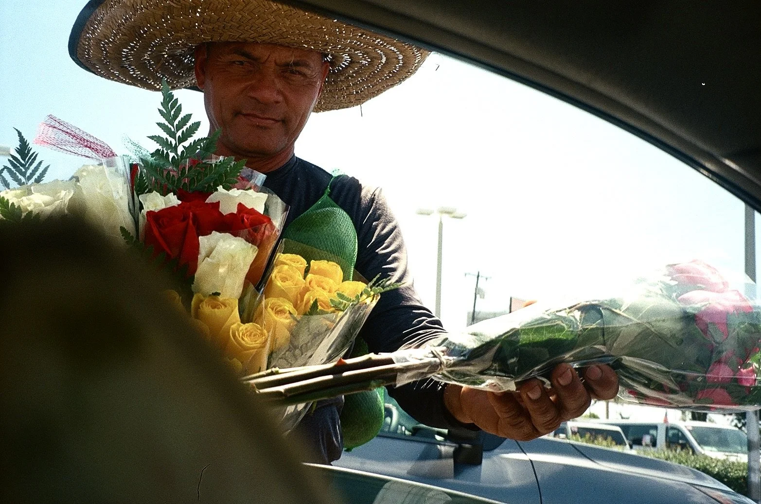 A man wearing a large straw hat is handing flowers through a car window. The flowers are wrapped in clear plastic and include roses in yellow, red, and pink, with some green leaves.