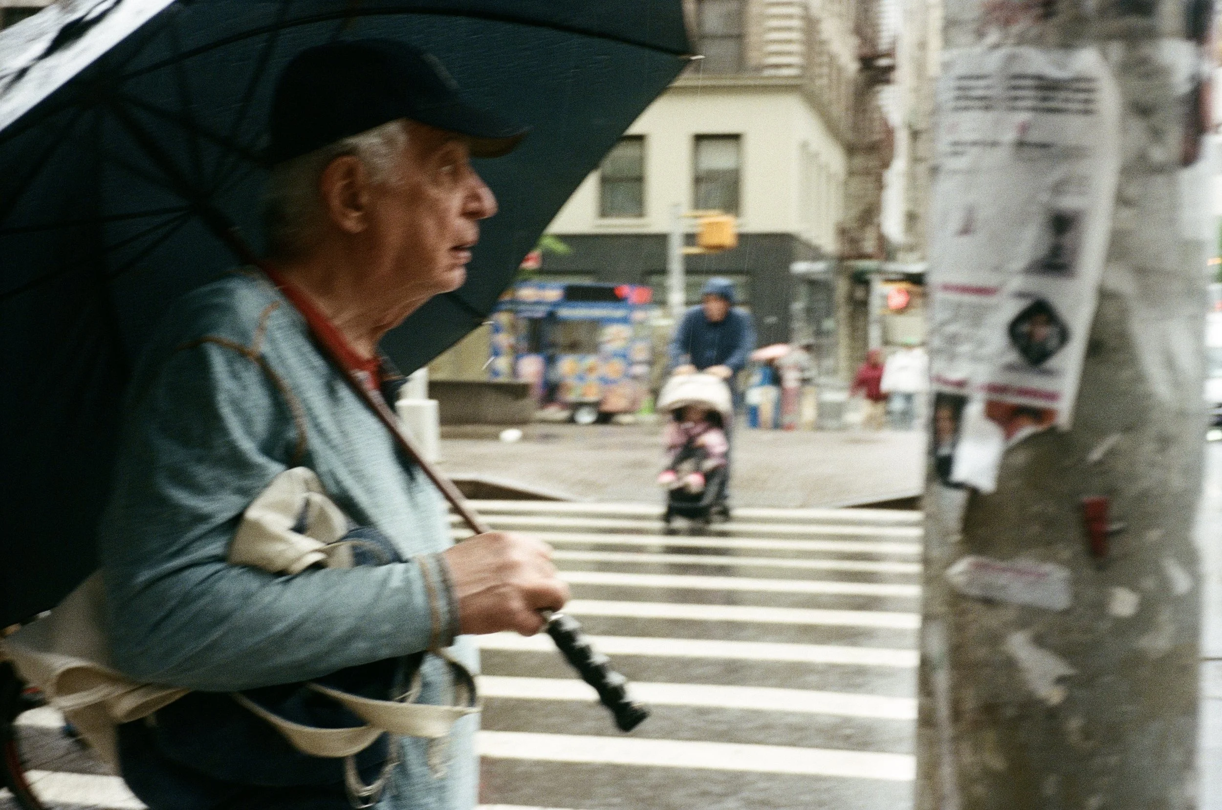 An elderly man with gray hair, wearing a black cap and a denim jacket, holding an umbrella and a striped walking cane, crosses a city street with a crosswalk on a rainy day.