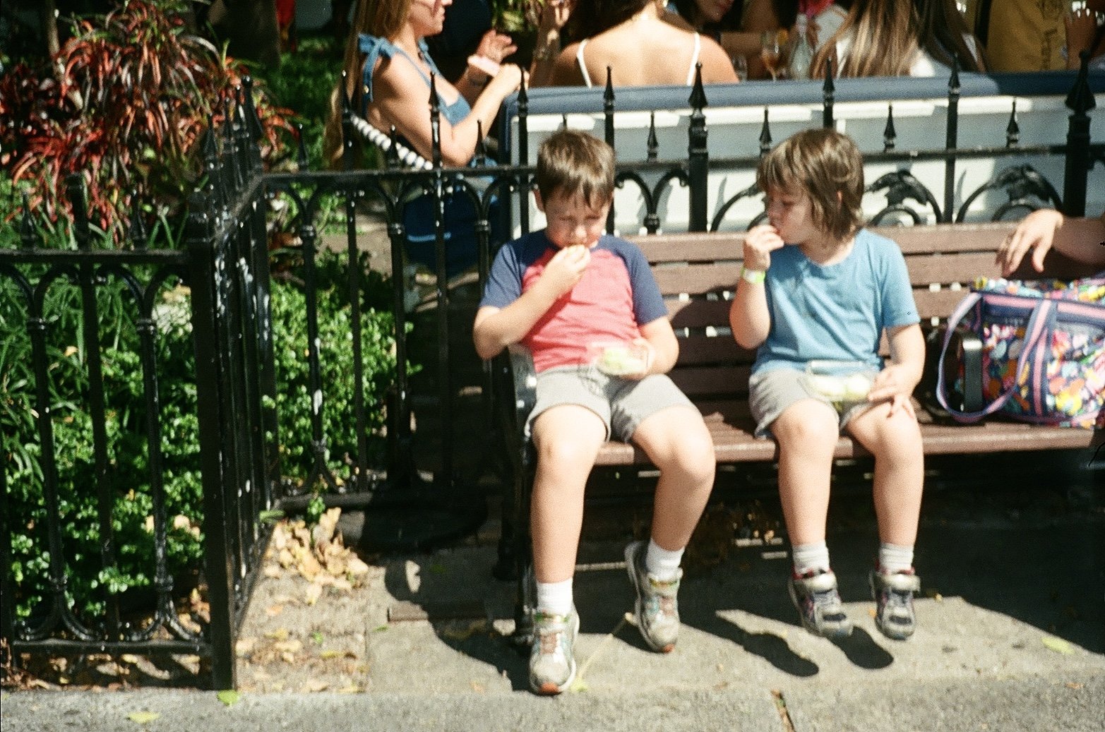 Two young boys sitting on a park bench eating ice cream. One boy is wearing a red and blue shirt with shorts, and the other is in a blue shirt with shorts. There are adults in the background, some standing and some sitting. The park is decorated with