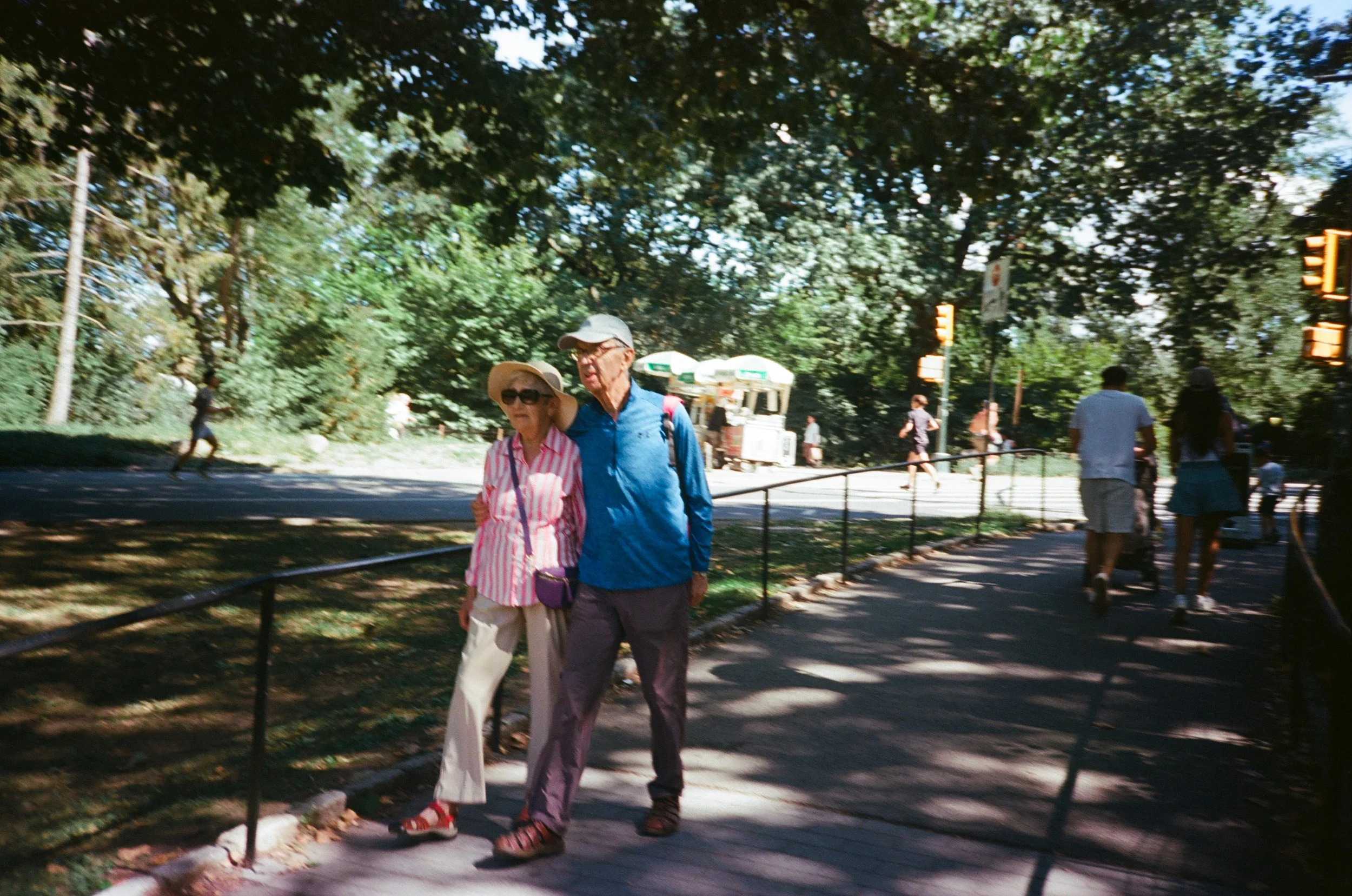 A couple walking along a park pathway surrounded by trees, with other people in the background.
