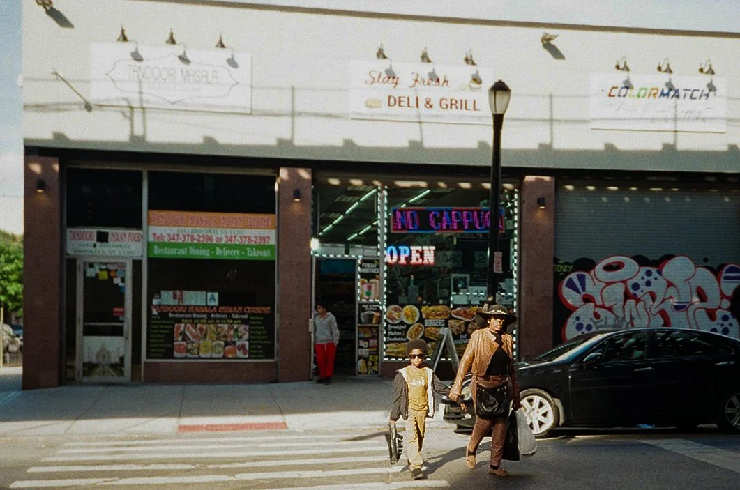 Street view of a storefront with signs reading 'Starry Fresh Deli & Grill' and 'Color Match.' People crossing the street in front, including a woman with a large sun hat and sunglasses holding a bag, a young boy with sunglasses and backpack, and a ma