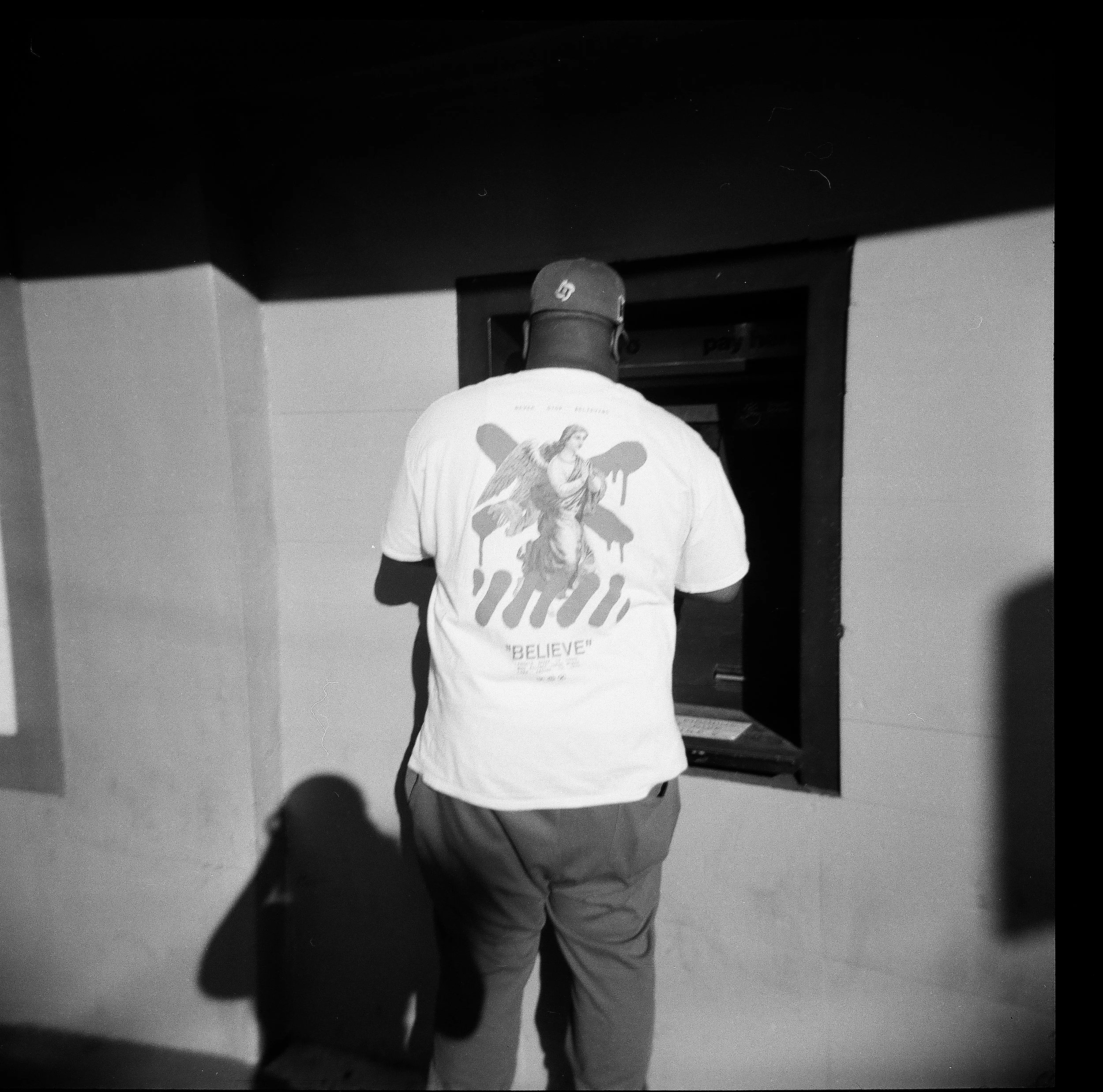 A man standing at an ATM, seen from behind, wearing a baseball cap and a white t-shirt with an angel graphic and the word 'BELIEVE' printed on it.