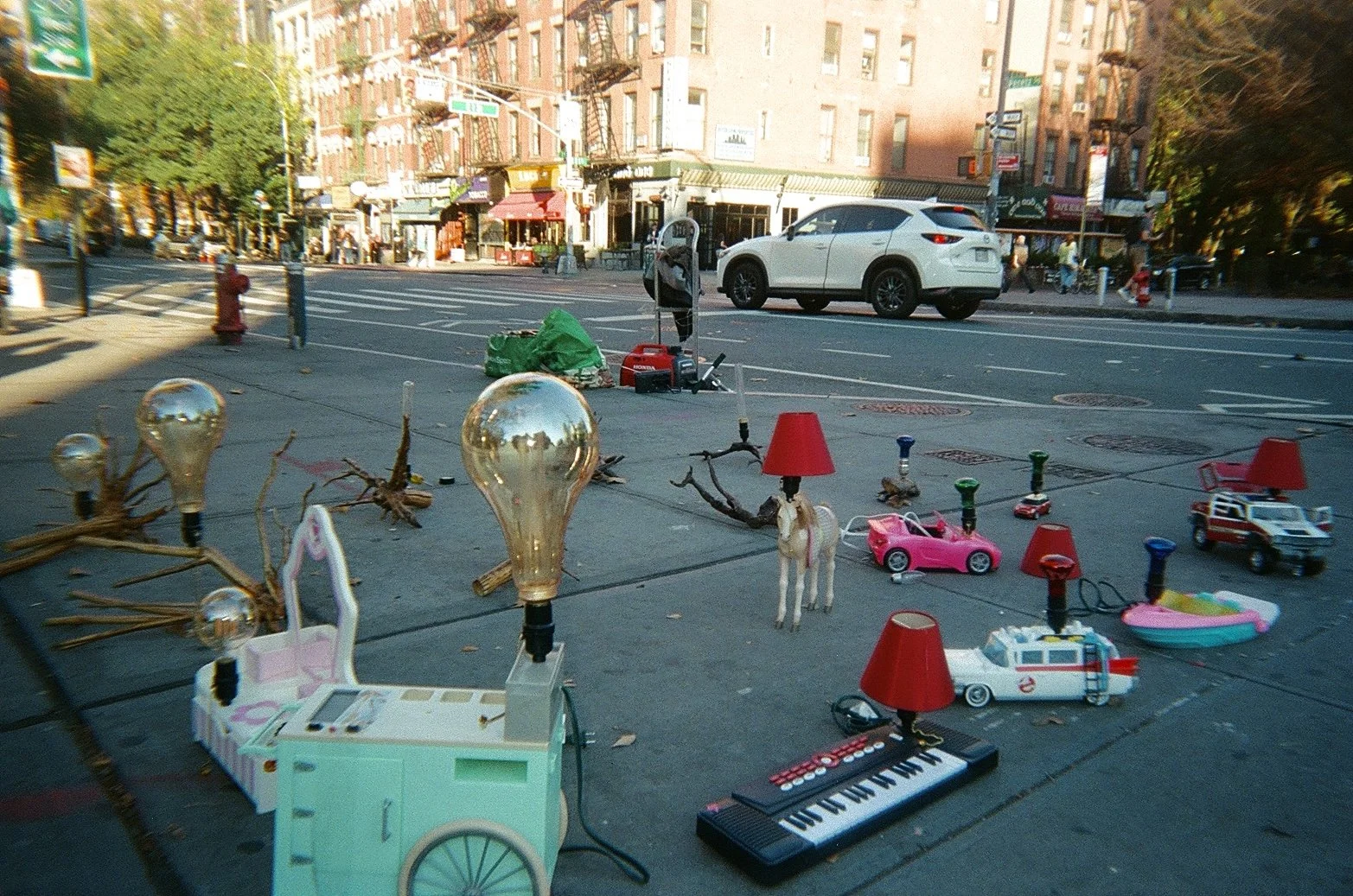 Street scene with eclectic street art and objects including light bulbs, a keyboard, several toy cars, a lamp with a horse head, branches, and other miscellaneous items on the sidewalk.