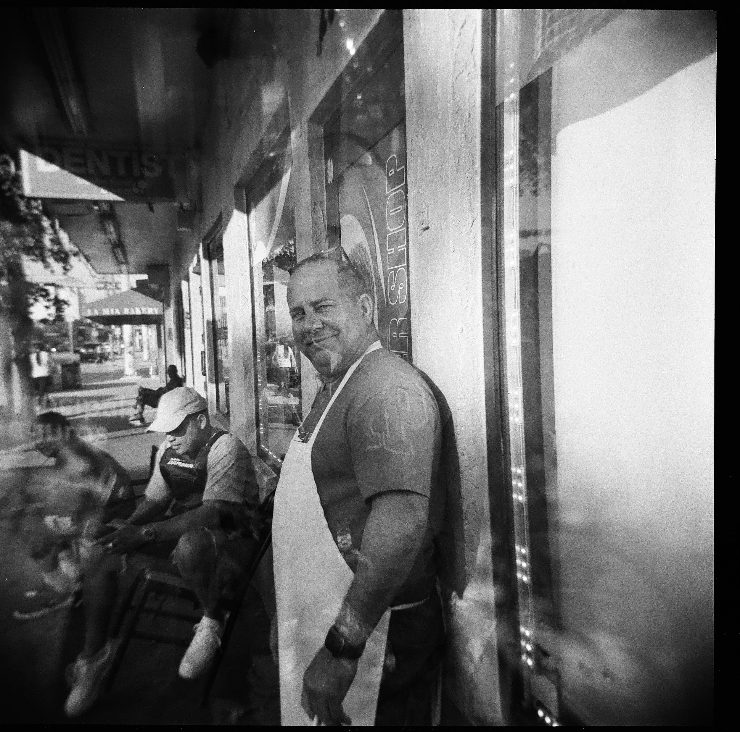Black and white photo of a man standing outside a store window, wearing a white apron and smiling. Two people are sitting by the window, one wearing a cap. The street scene with cars and buildings is visible reflected in the window.