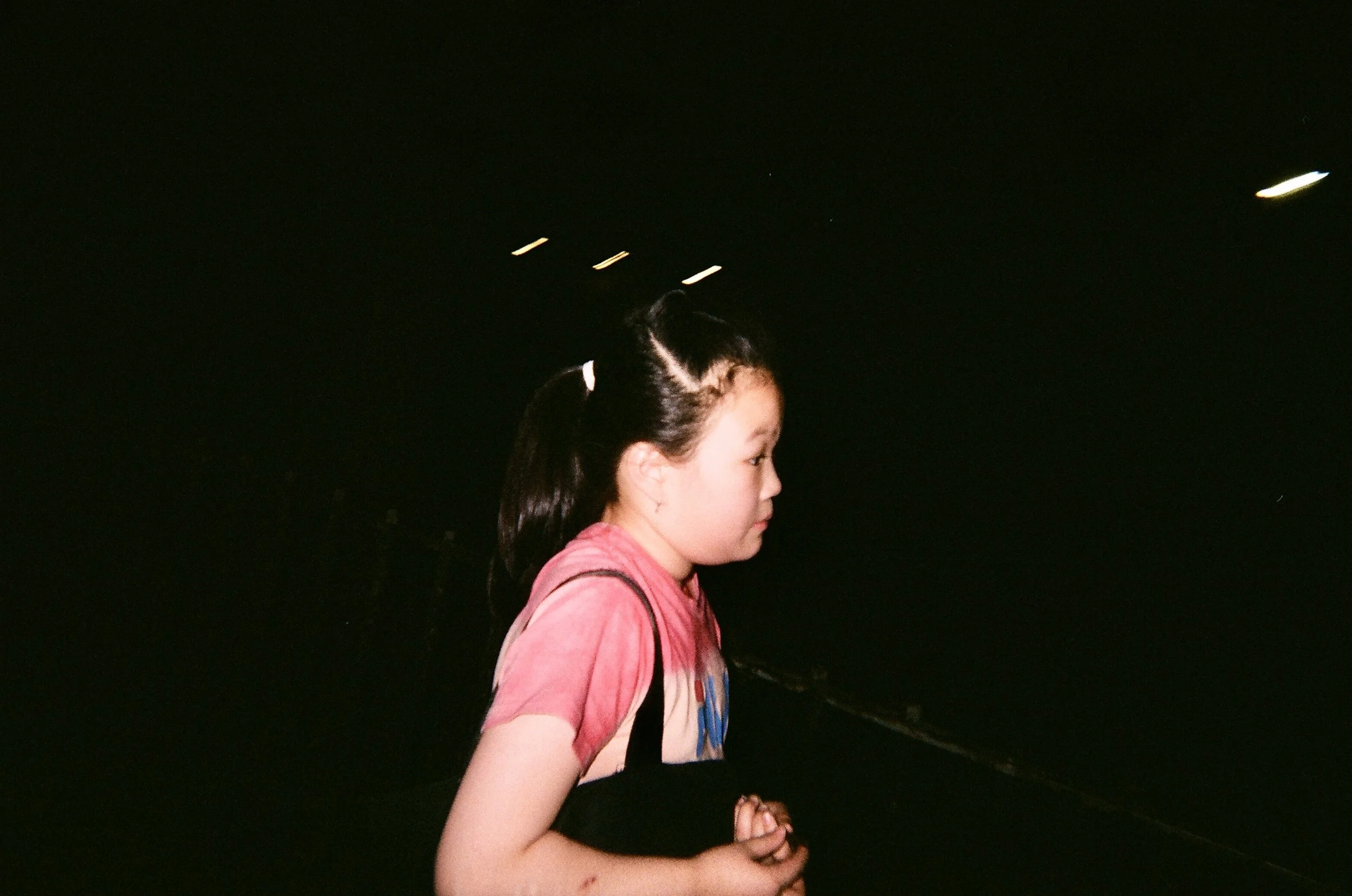 A young girl with dark hair in pigtails, wearing a pink tie-dye shirt, with her hands clasped, walking outdoors at night.