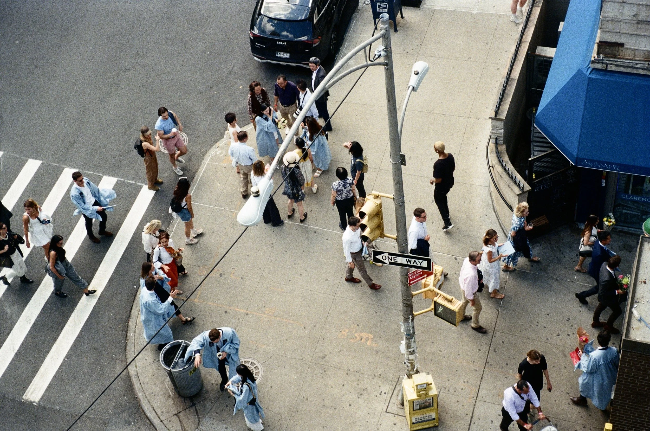 Bird's eye view of people on a city sidewalk, some wearing graduation gowns, walking in various directions near a crosswalk, streetlight, and parking lot, with cars parked nearby.