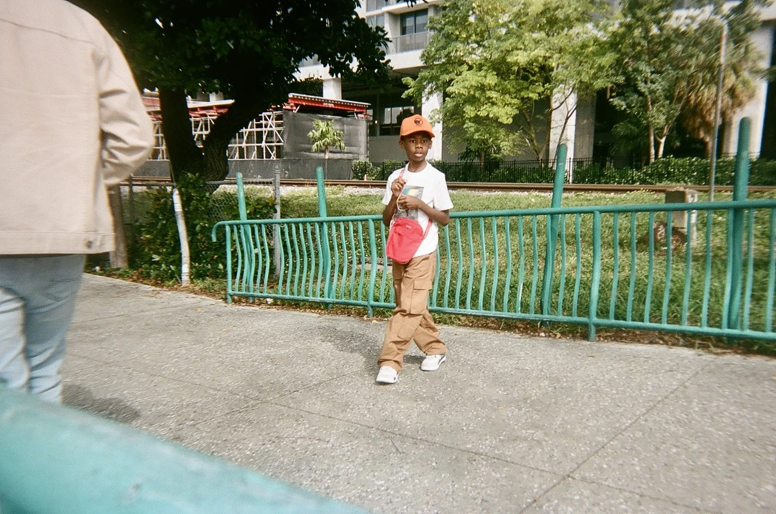 A young boy wearing a white t-shirt, khaki pants, white sneakers, and an orange cap walking along a sidewalk near a green metal fence and trees in an urban park setting.