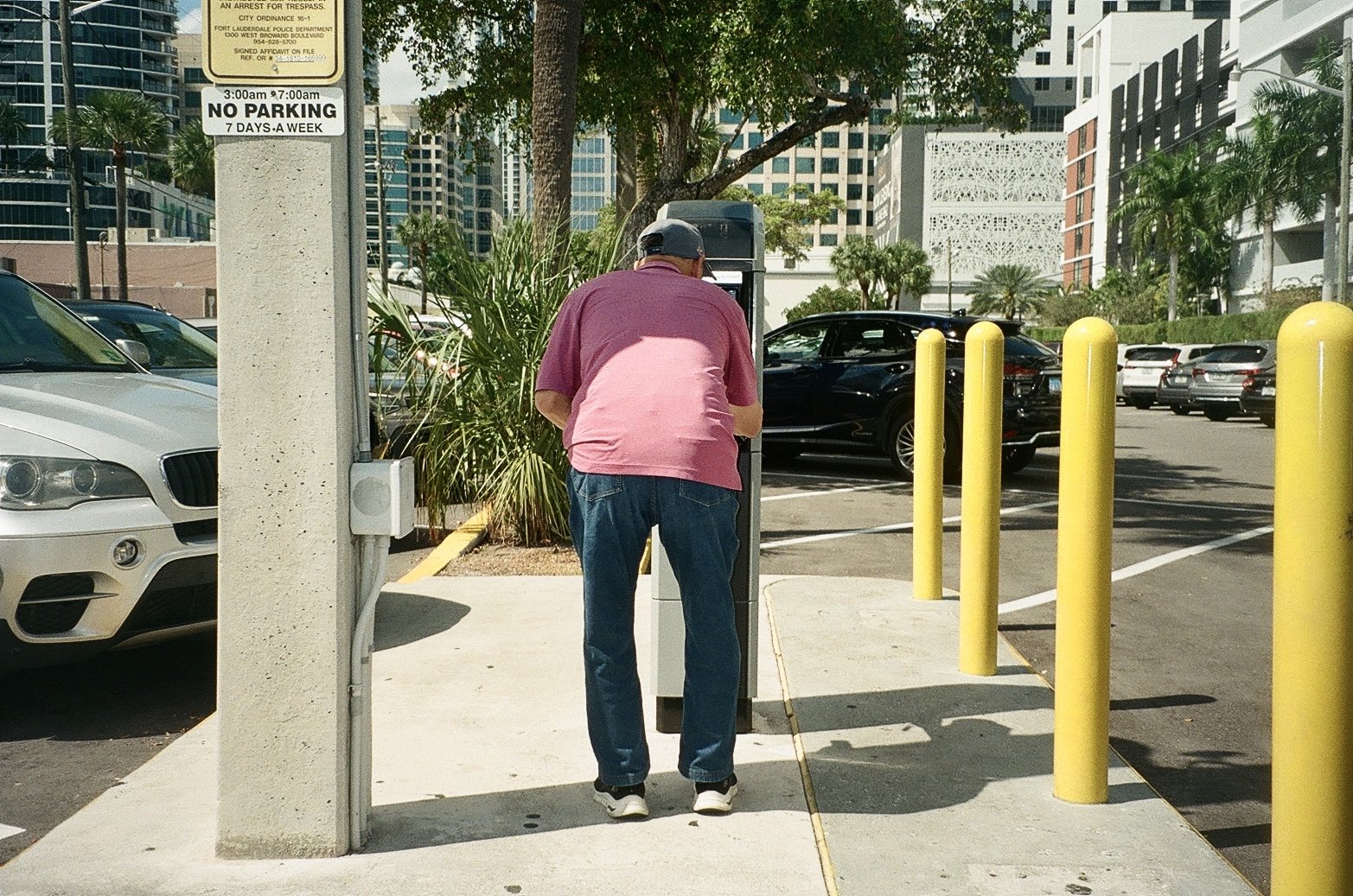 A man in a pink and burgundy shirt and jeans is using a payphone on a sidewalk in a parking lot, with yellow posts, parked cars, trees, and tall buildings in the background.