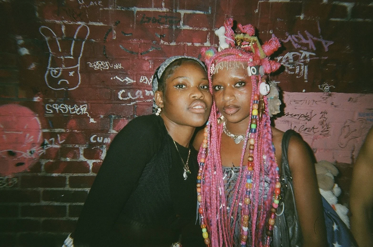 Two women with distinctive hairstyles and accessories stands close together in front of a graffiti-covered brick wall. One woman has grayish hair and loops earrings, the other has bright pink dreadlocks adorned with beads and various accessories. The