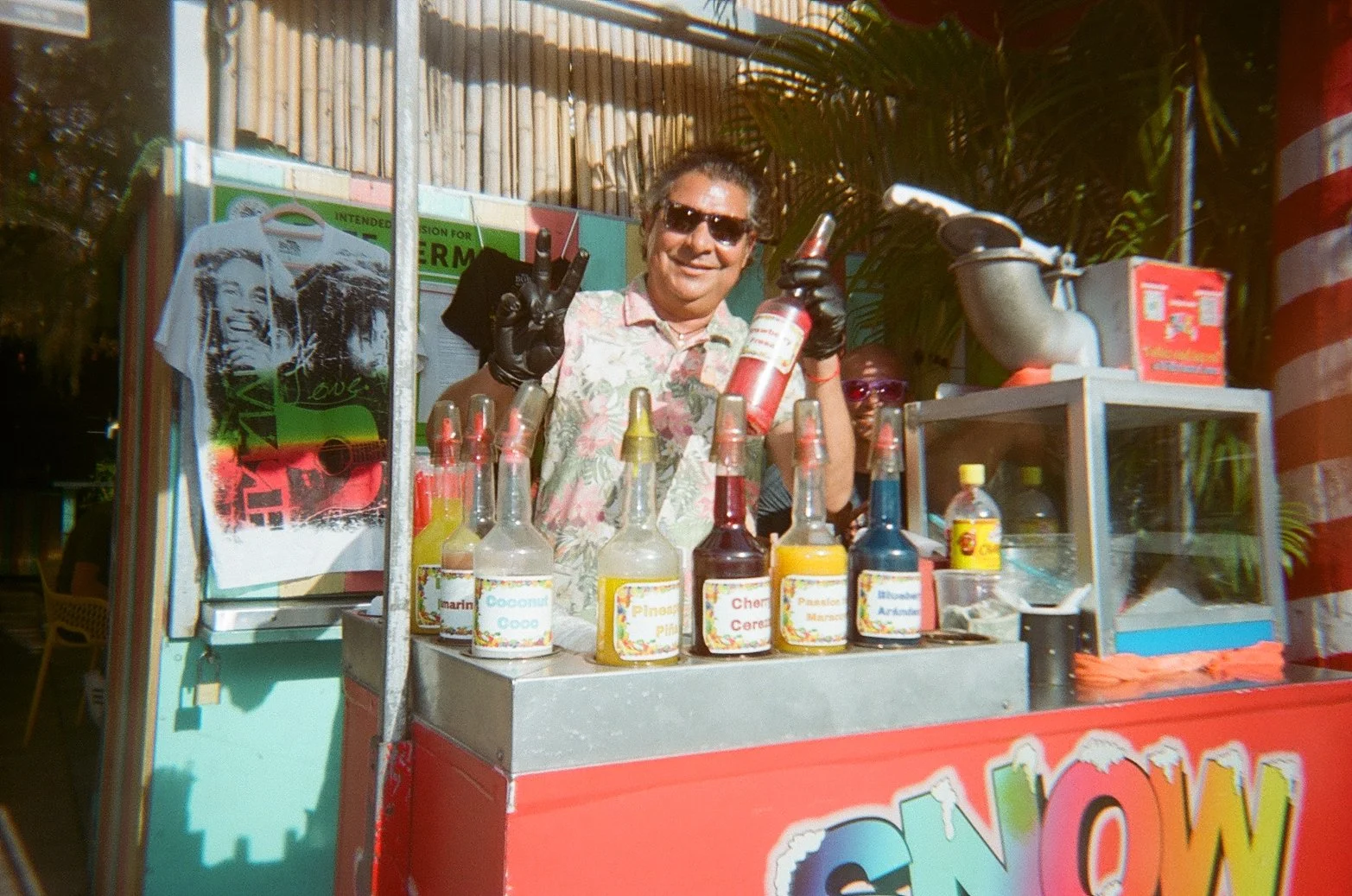 A man with sunglasses and a floral shirt standing behind a colorful street drink cart, holding a fire extinguisher and making a peace sign. The cart has various bottles of flavored syrups and a poster of a young girl with the word 'WOW' on a red back