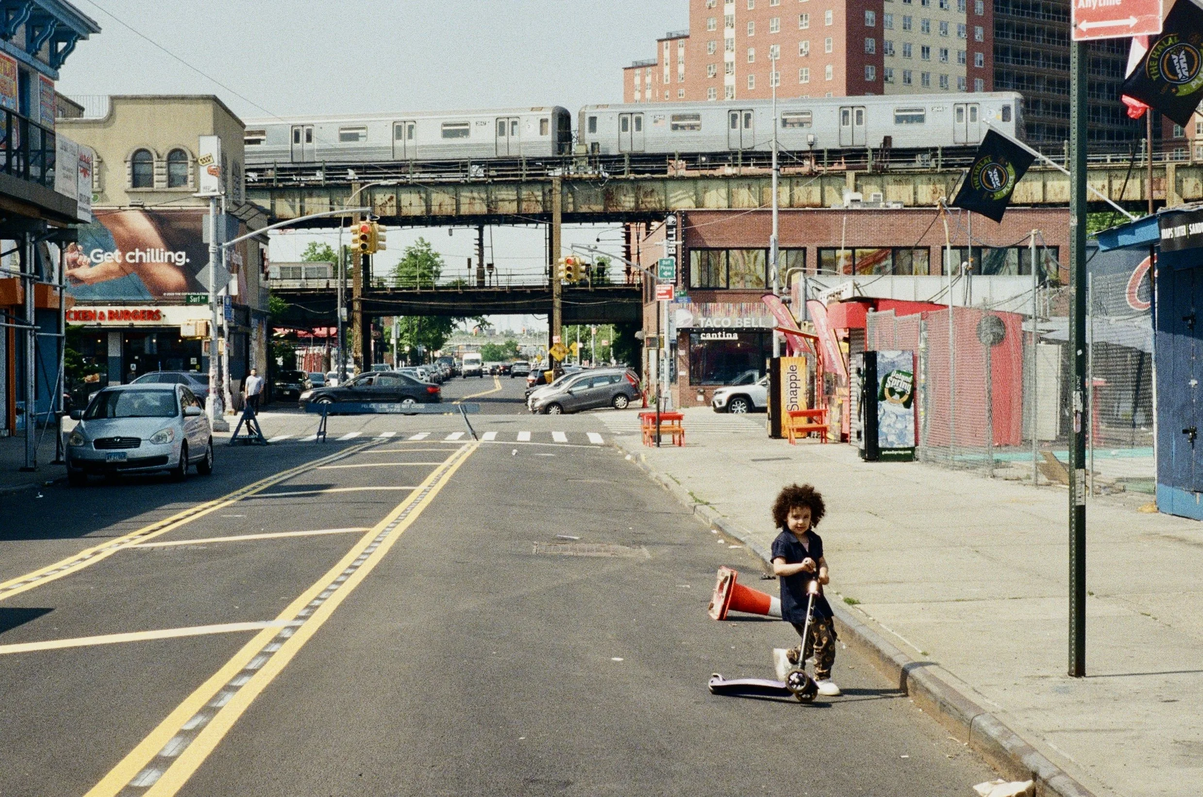 A young girl with curly hair stands on an urban street corner, holding onto a scooter. There are parked cars, a Taylor Swift billboard, and a train traveling on elevated tracks in the background. Bright daylight and city buildings are visible.