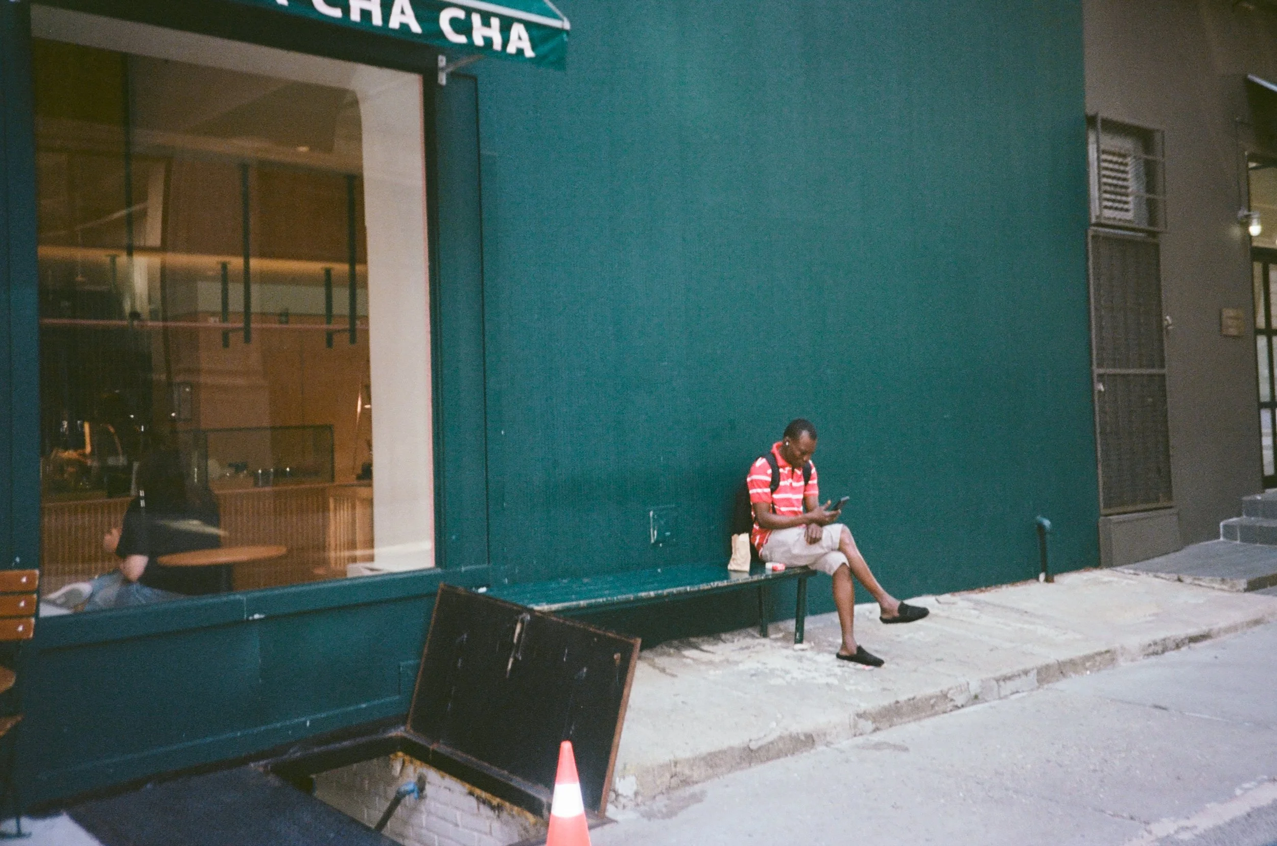 A man sitting on a bench outside a green building, wearing a red and white striped shirt, khaki shorts, and black slippers, looking at his phone.