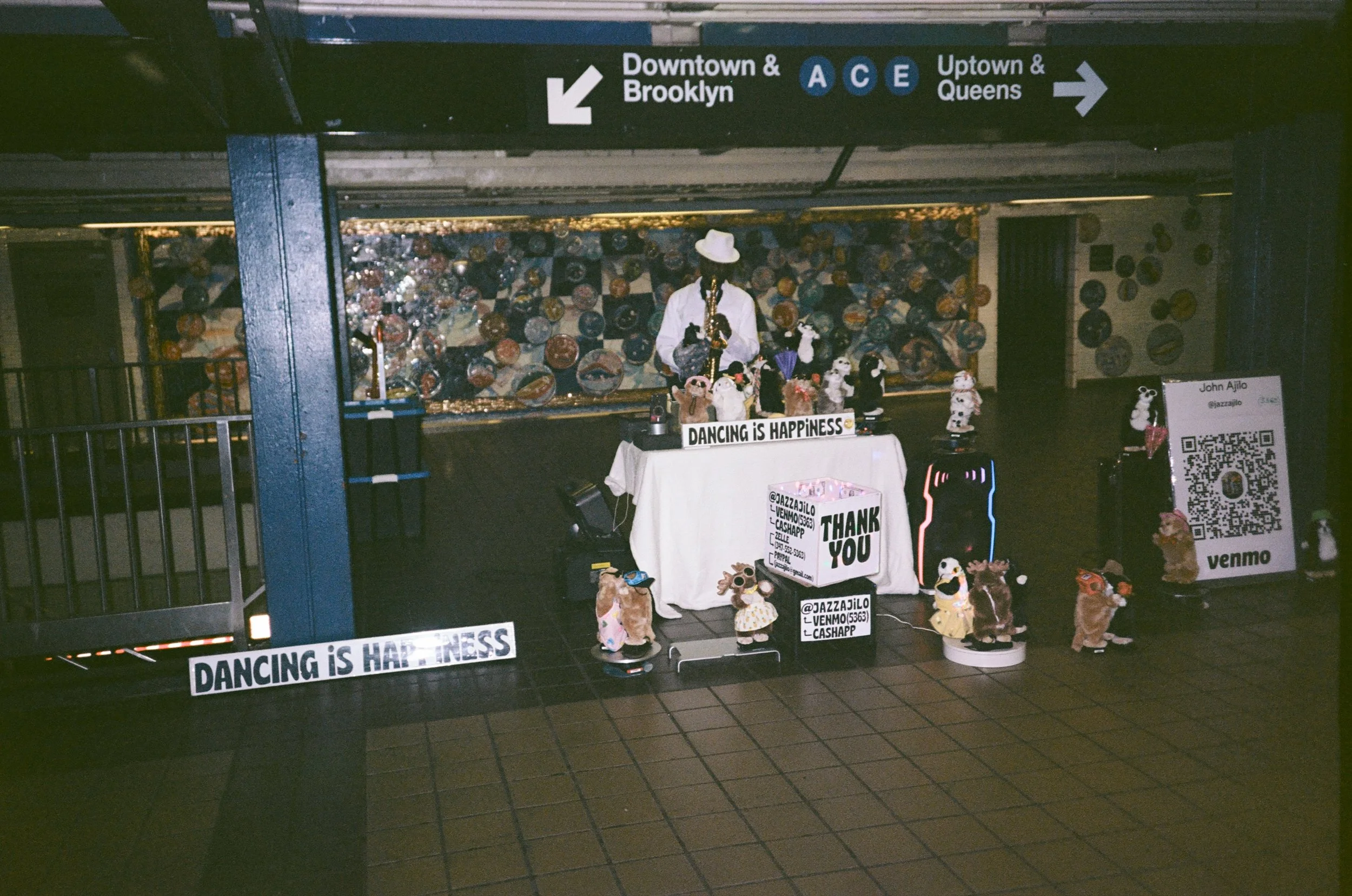 Underground subway station with a small display of handmade stuffed animals and signs. Signs say 'Dancing is Happiness' and 'Thank You.' There is a QR code for Venmo. The display is behind a black railing with entry/exit signs pointing towards Brookl