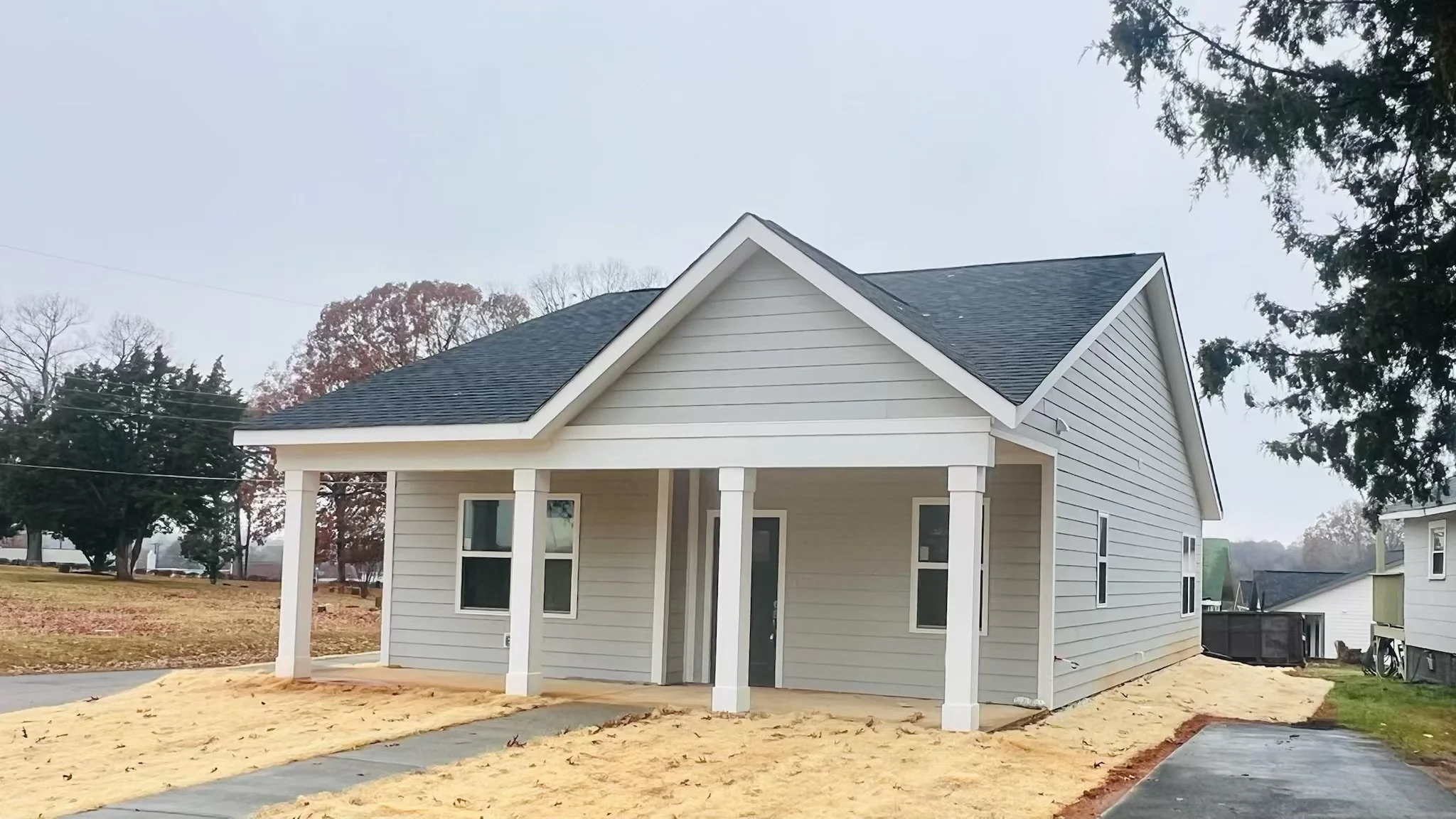 Newly constructed light gray house with white trim, a dark gray roof, and a front porch supported by four white columns, with a concrete walkway leading to the front door, surrounded by a sandy lawn and neighboring houses.