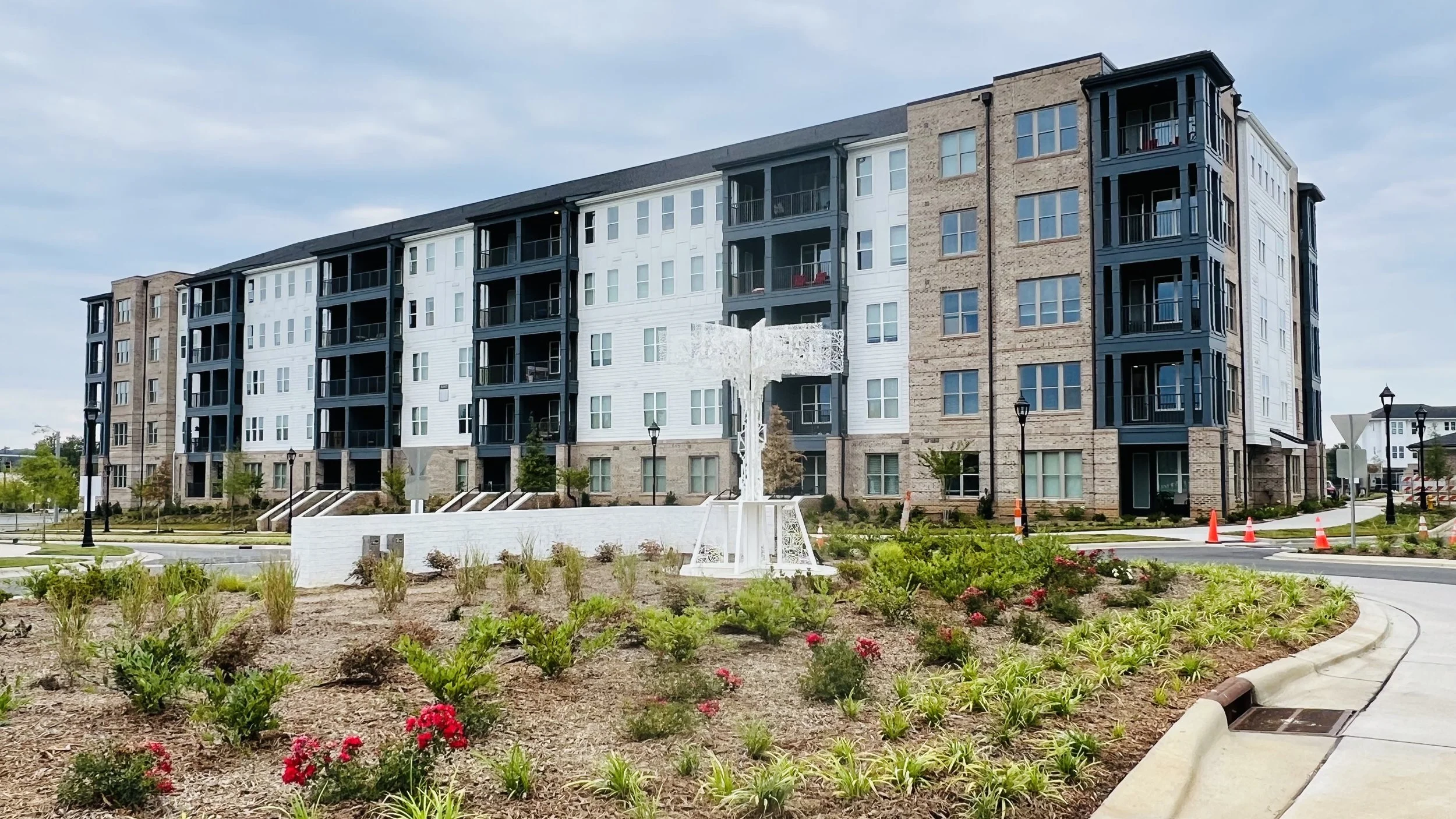 A modern multi-story apartment building with a landscaped front yard and a white sculpture resembling a tree. The building features a mix of brick and white siding, with multiple balconies, and the scene includes landscaping