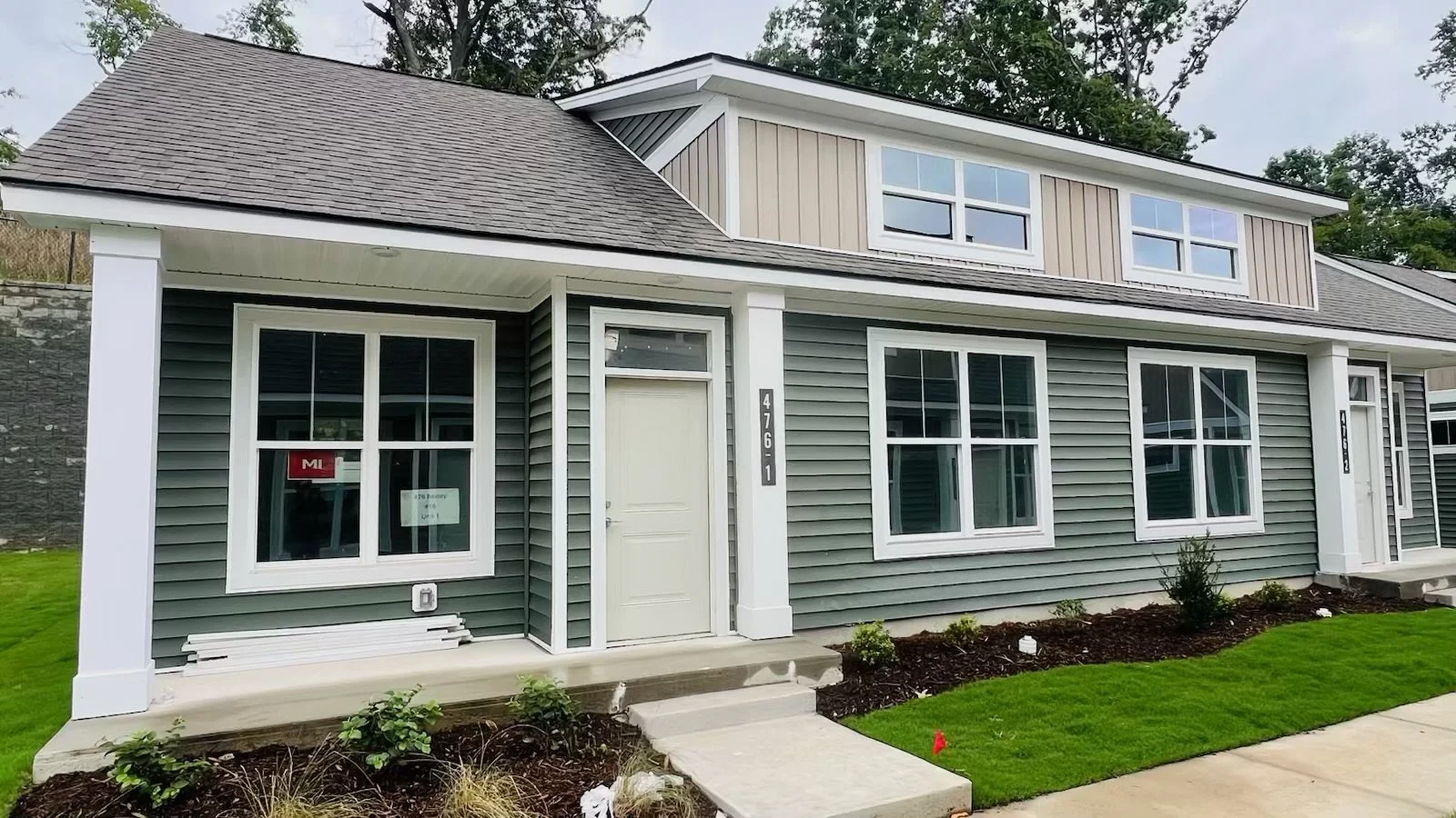 Newly built two-story house with gray siding, white trim, and multiple large windows, surrounded by a manicured lawn and flower bed.