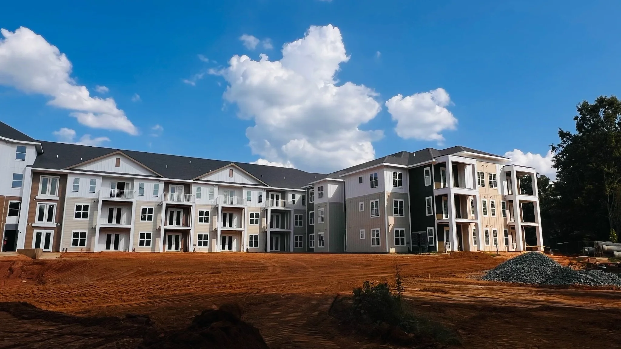 New multi-story apartment complex under construction with a dirt foreground and a clear blue sky with clouds in the background.