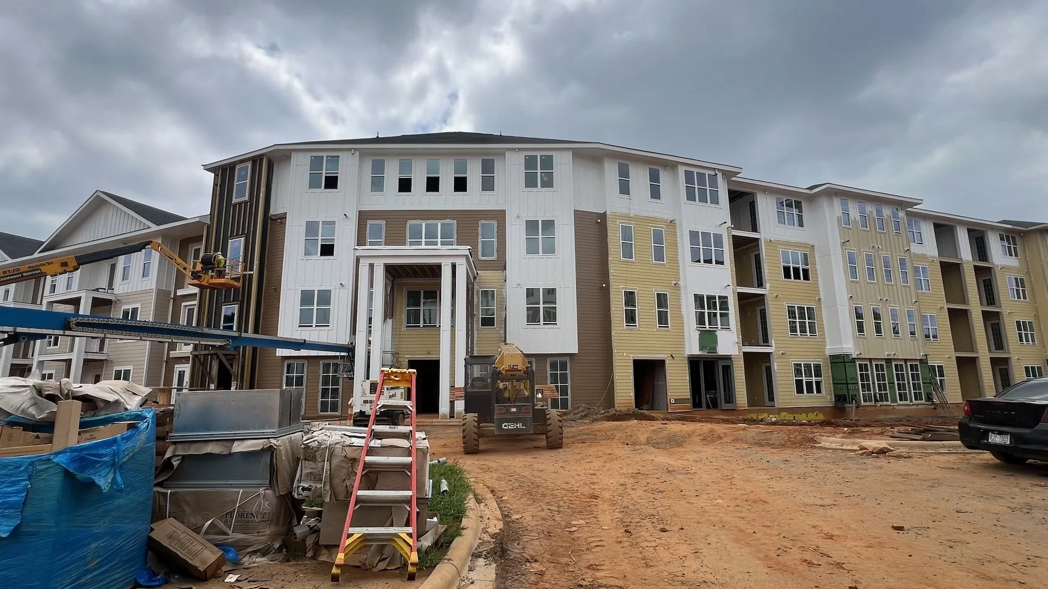 Under construction apartment building with scaffolding and construction equipment, dirt driveway, and cloudy sky. Made of Brick and siding