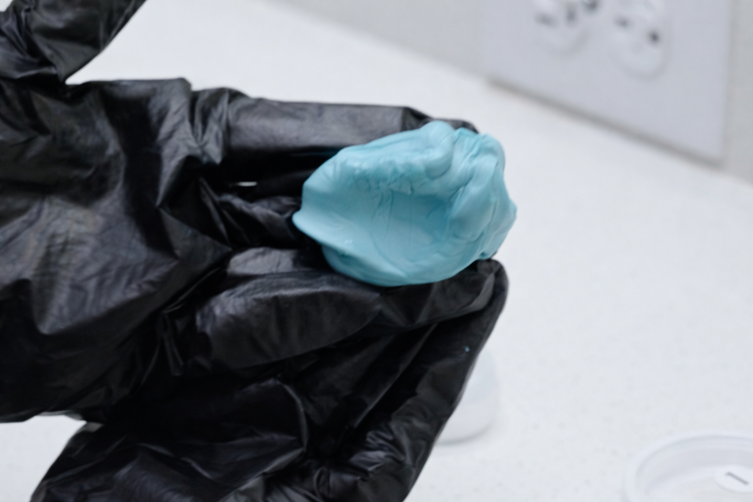 A person's hand wearing a black glove holding a blue substance in a small, irregularly-shaped blob.