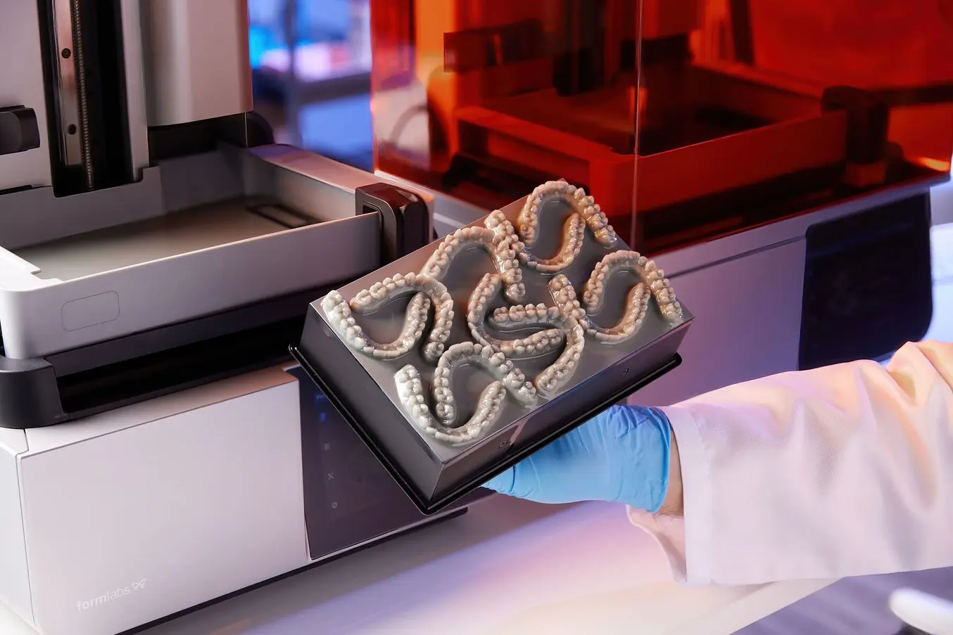 A scientist wearing a lab coat and blue gloves placing a tray with multiple 3D printed teeth on a laboratory device in a lab setting.