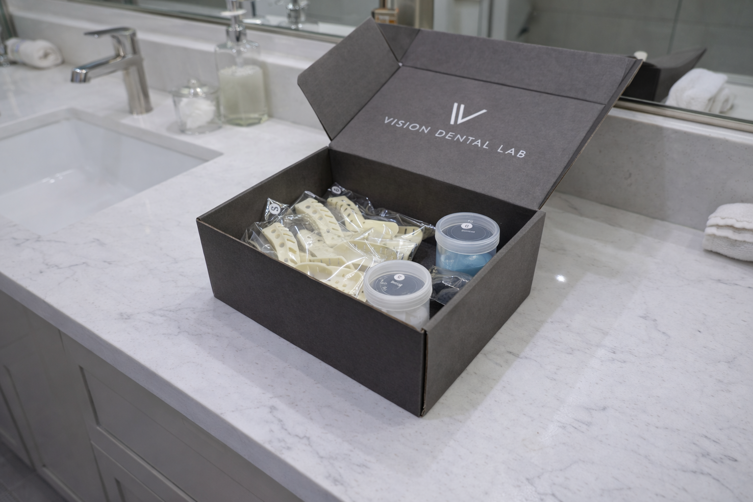 Open black box labeled 'Vision Dental Lab' containing dental impression trays and small containers, placed on a white marble bathroom countertop near a sink with soap dispensers and towels.