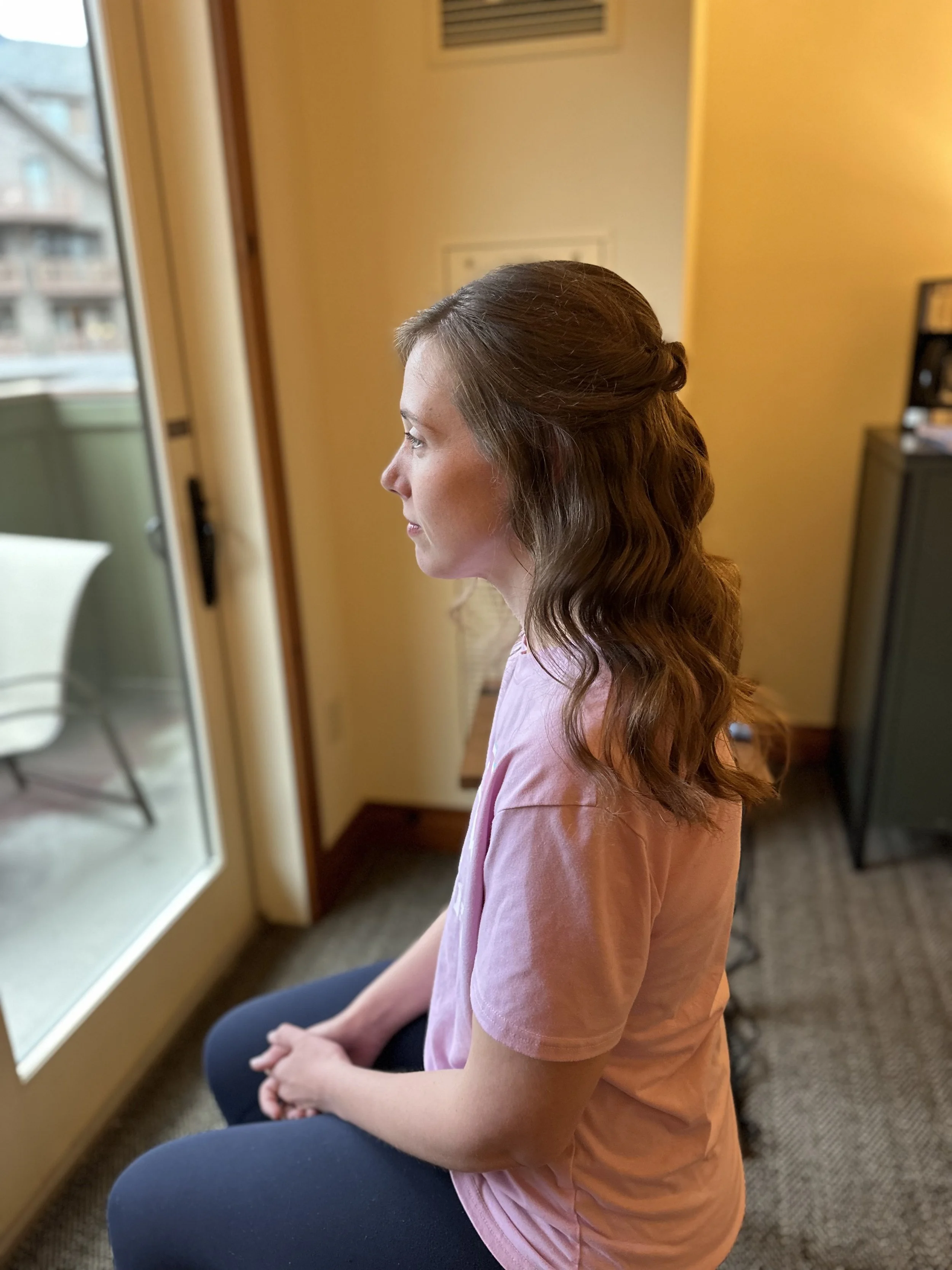 A Bride to Be. Hair Trail in Stowe Vermont. She has wavy brown hair is sitting indoors near a glass door, looking to the left, with her hands clasped in her lap. She is wearing a pink t-shirt and dark pants.