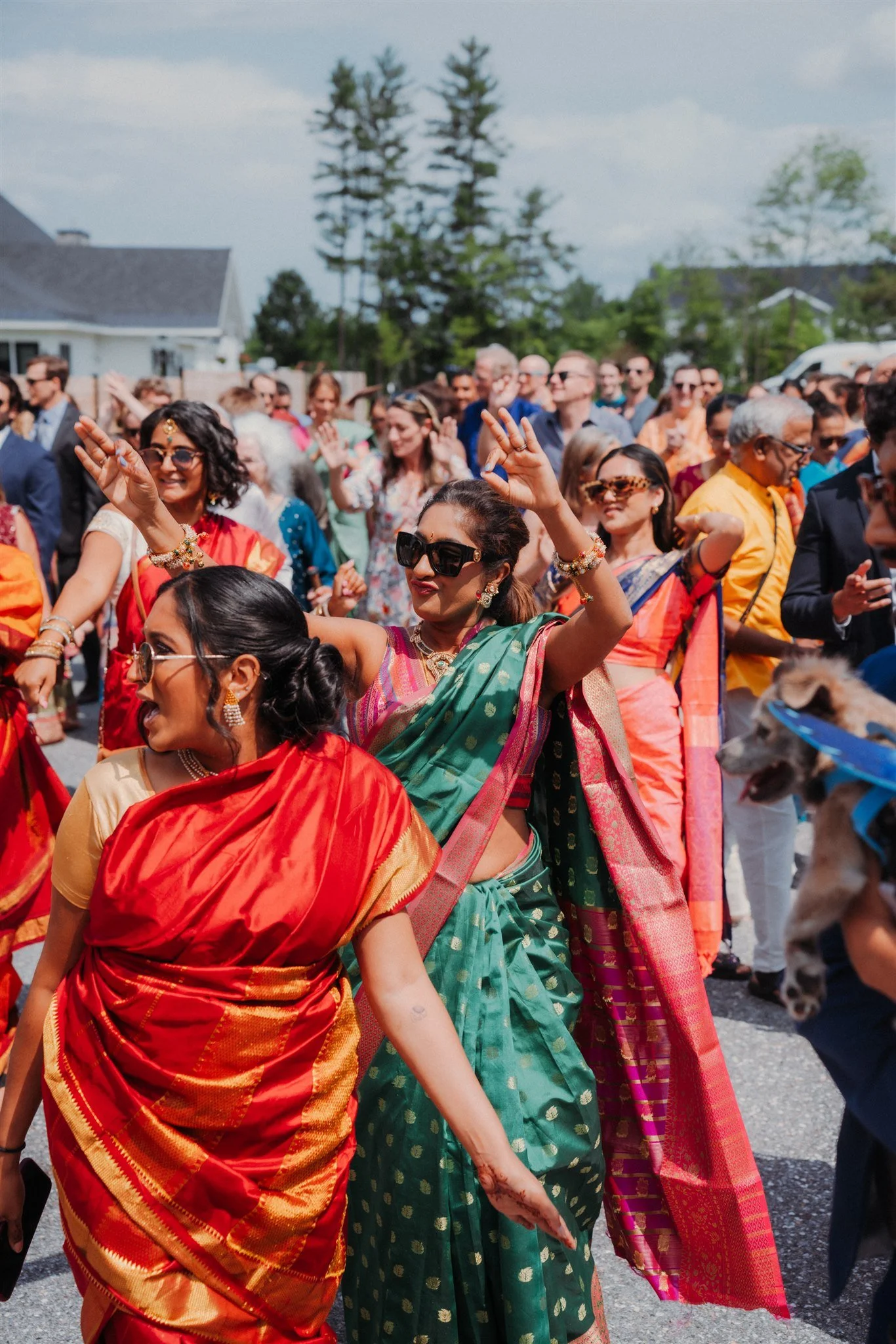 People dancing outdoors at a celebration, dressed in traditional Indian sarees and colorful attire, with some wearing sunglasses, under a partly cloudy sky at the Essex Resort & Spa Vermont