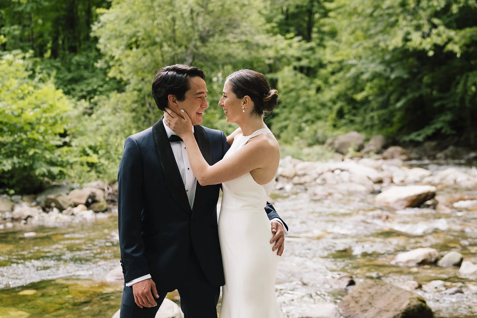 Bride and Groom. A couple dressed in wedding attire stands in a lush green outdoor setting near a rocky stream, smiling and looking into each other's eyes in Bristol, Vermont