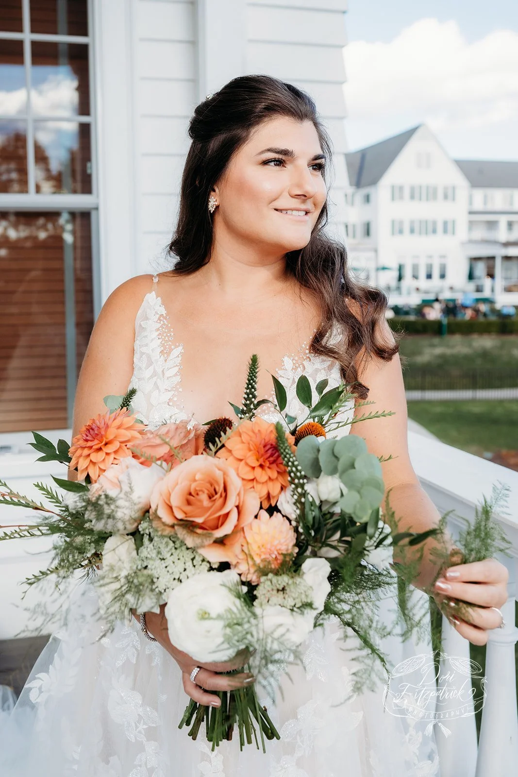 A bride with dark brown hair in loose curls, wearing a white lace wedding dress, holding a large bouquet of peach, white, and green flowers, standing outside on a porch with residential buildings in the background. Vermont Bridal Hairstyle. Wildflowe