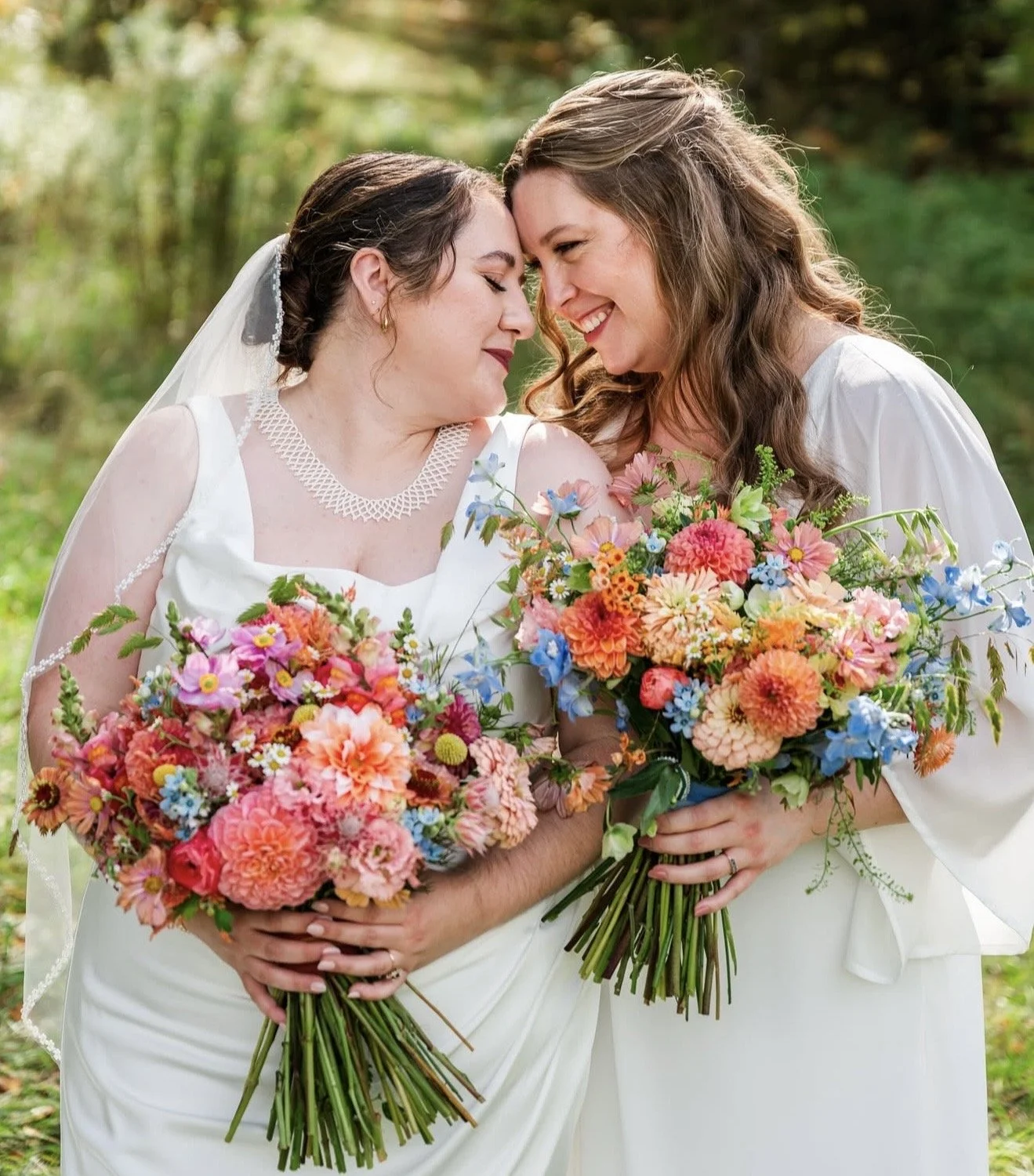 Two women in white dresses holding large bouquets of colorful flowers, touching foreheads, smiling in an outdoor setting with greenery. Vermont Bridal Hairstyle. Wildflower Beauty Company. Burlington VT, Stowe, Manchester VT. Vermont Wedding, Vermont