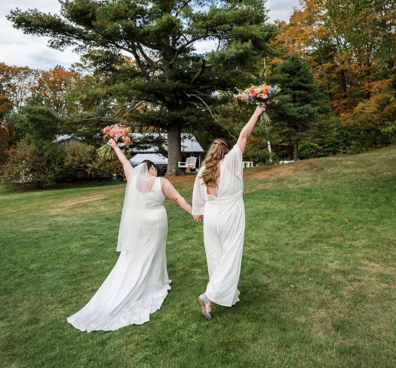 Two brides in white wedding dresses holding wedding bouquets, walking hand in hand outdoors on grassy lawn with trees and a house in the background. Vermont Bridal Hairstyle. Wildflower Beauty Company. Burlington VT, Stowe, Manchester VT. Vermont Wed