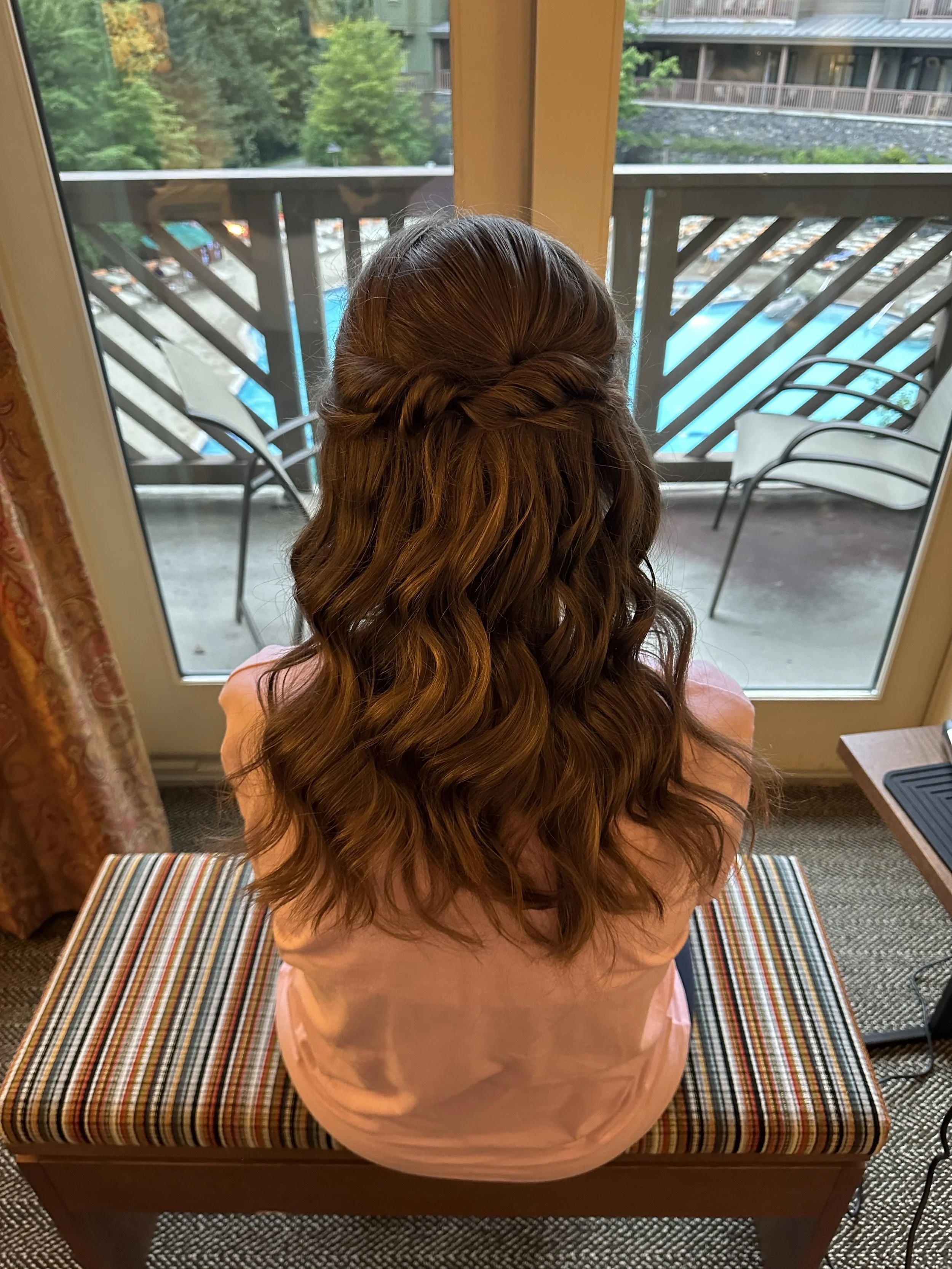 A woman with long, wavy, brown hair sitting on a striped cushioned bench, facing a glass door that opens to a balcony with a swimming pool and outdoor chairs, surrounded by greenery.
