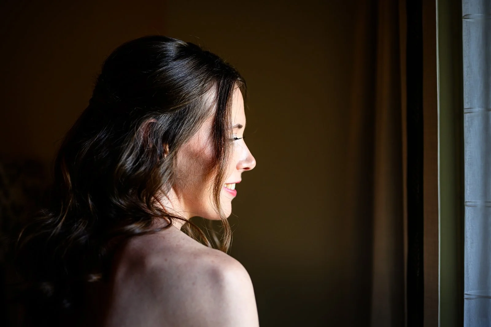 Profile of a smiling woman with dark wavy hair looking out a window. Vermont Bridal Hairstyle. Wildflower Beauty Company. Burlington VT, Stowe, Manchester VT. Vermont Wedding, Vermont Bride, Vermont Wedding Hair