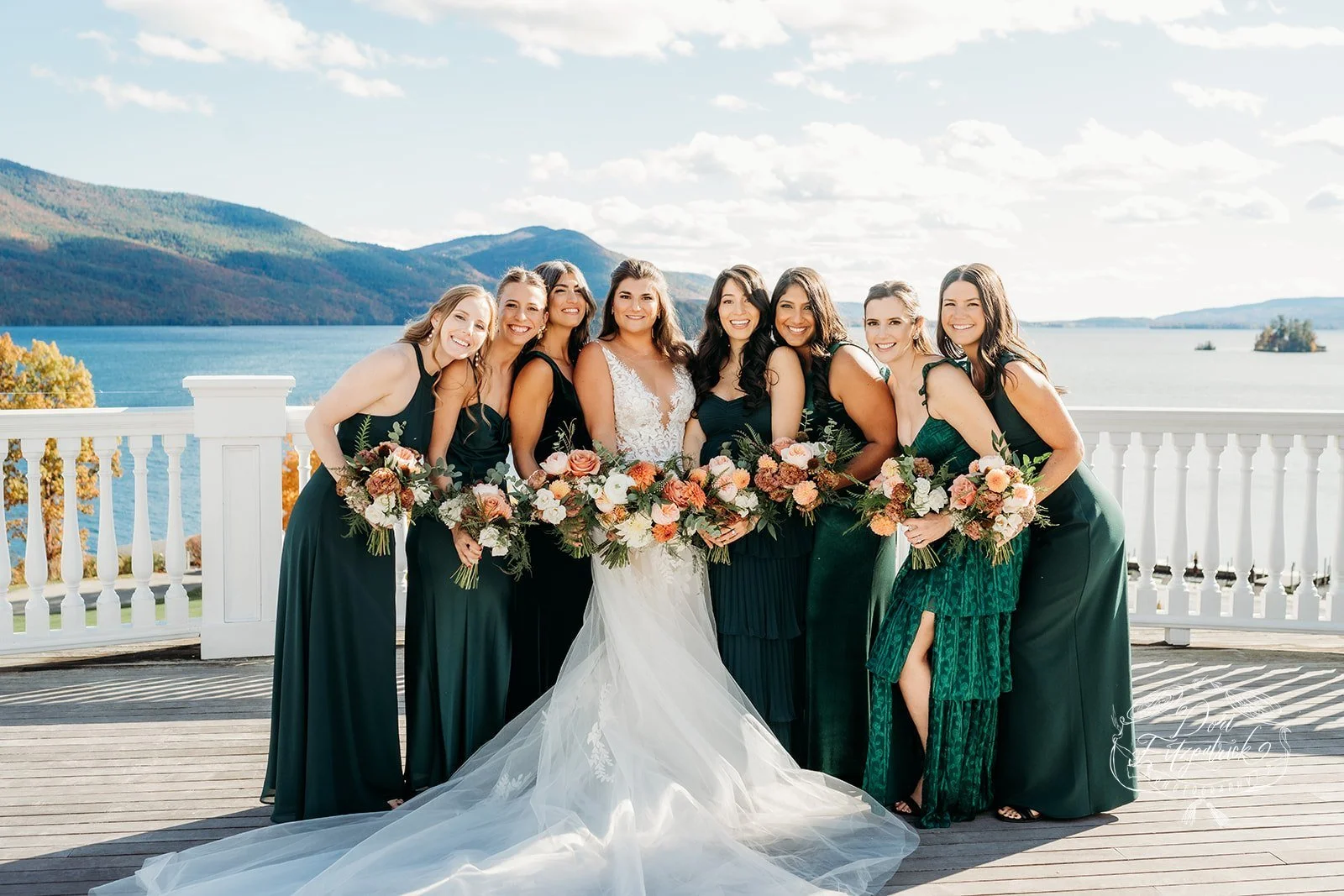 A bride in a white gown with a long train and eight bridesmaids in dark green dresses standing on a wooden deck by a white railing, with a lake, mountains, and blue sky in the background, during a wedding celebration.