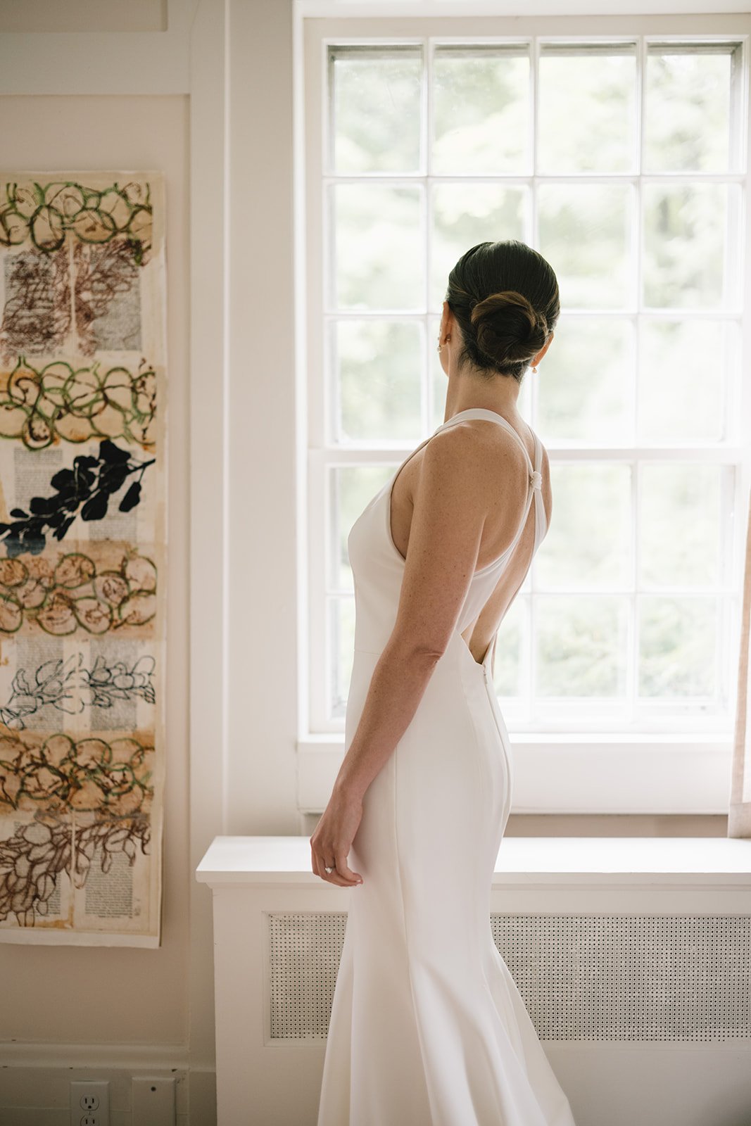A woman in a white wedding dress standing by a window, viewed from the back. Vermont Bridal Hairstyle. Wildflower Beauty Company. Burlington VT, Stowe, Manchester VT. Vermont Wedding, Vermont Bride, Vermont Wedding Hair