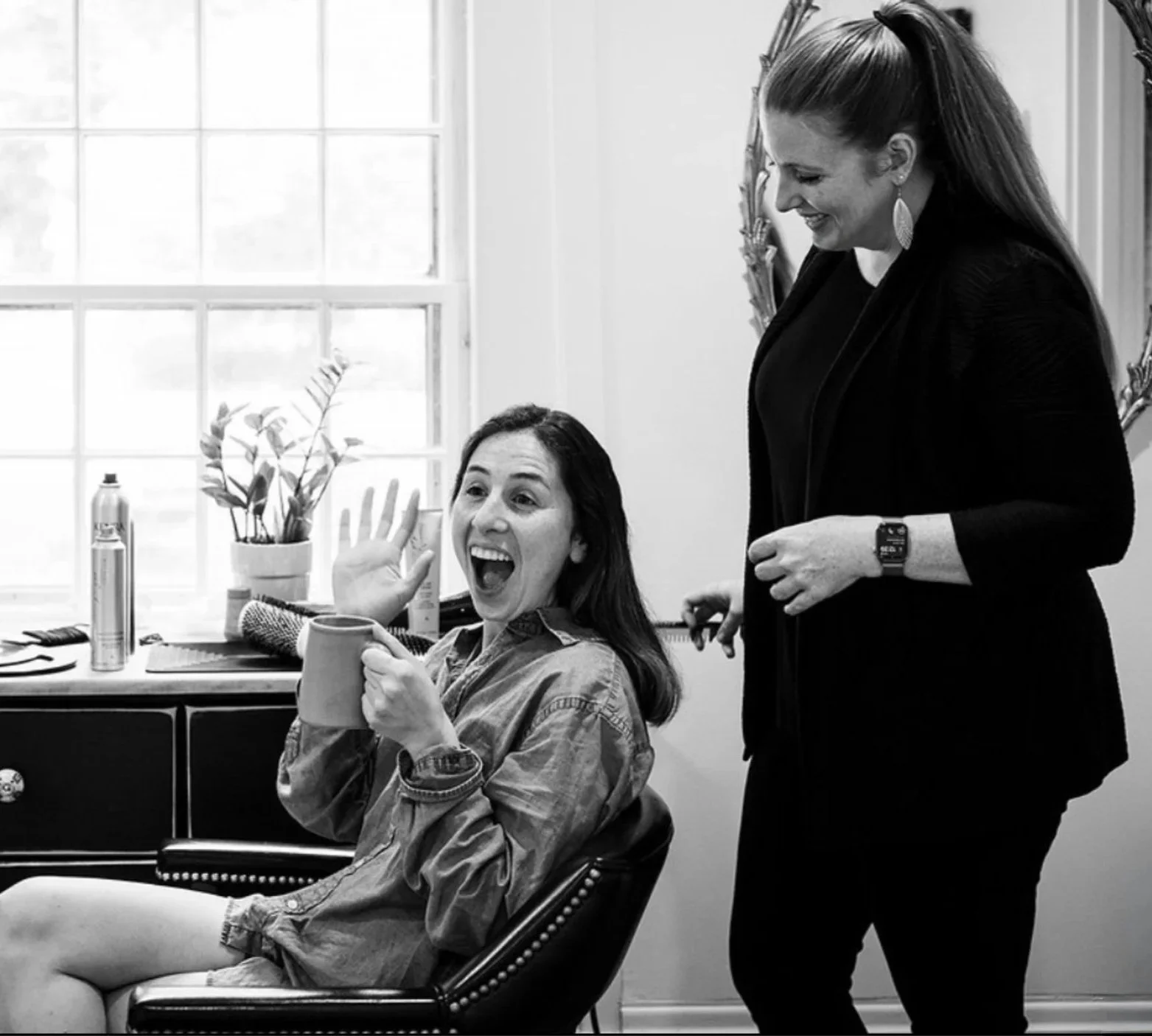 Megan Cole styling hair for the bride to be. Bride is sitting in a chair holding a mug, smiling and waving, the other standing and smiling, dressed in black at The Tillerman Inn Bristol Vermont