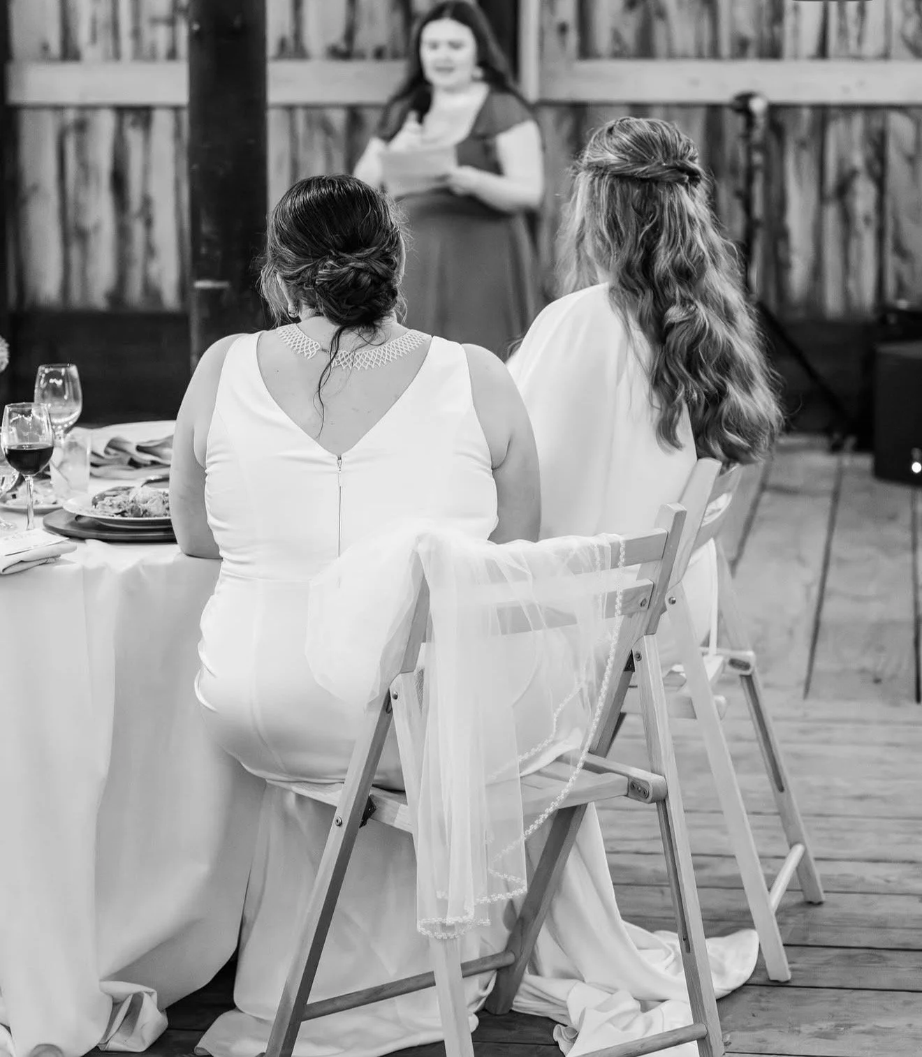 Two women sitting at a table during a wedding reception, viewed from the back, listening to a woman speaking in front of a wooden wall. Vermont Bridal Hairstyle. Wildflower Beauty Company. Burlington VT, Stowe, Manchester VT. Vermont Wedding, Vermont